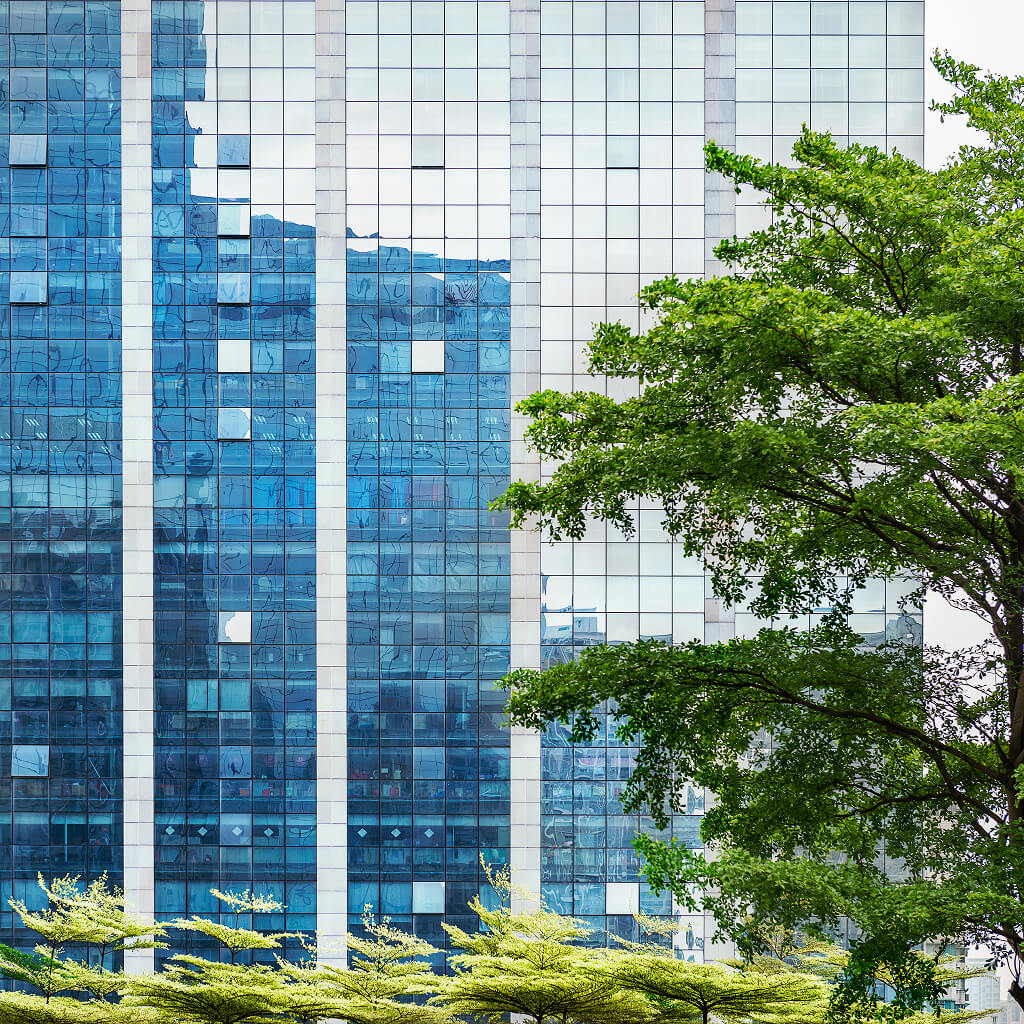 Glass skyscraper with reflective blue windows next to green trees and foliage in an urban setting.
