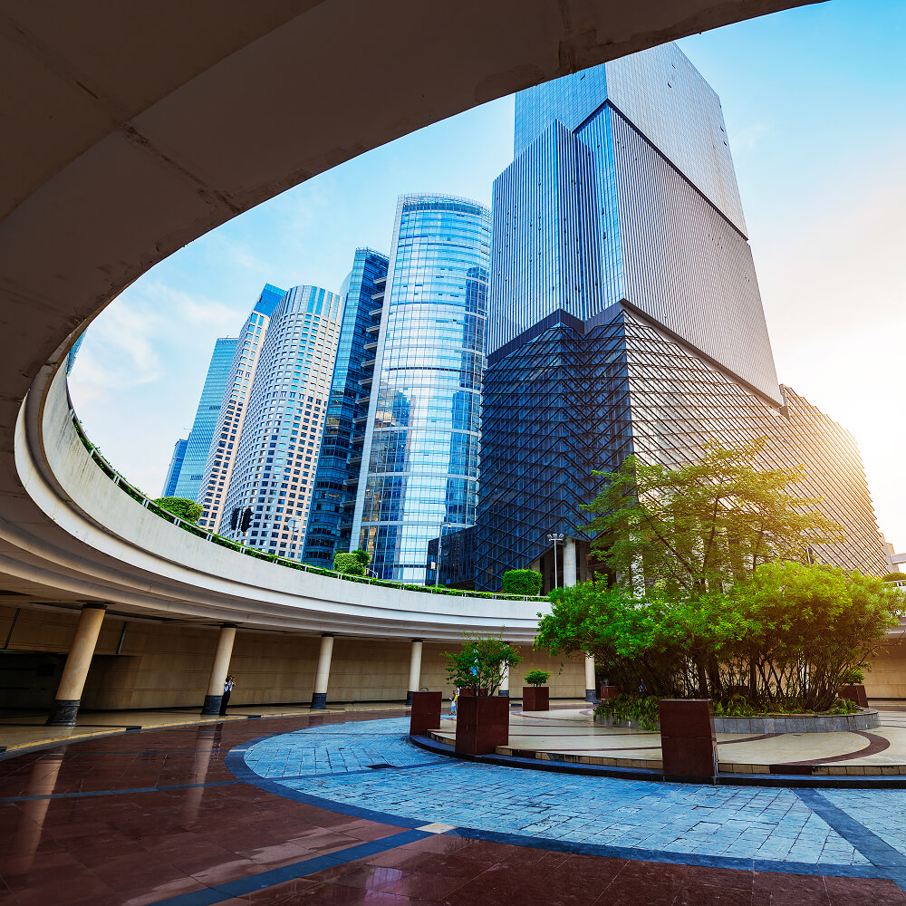 Modern skyscrapers behind a curved open plaza with trees and tiled ground in the foreground.