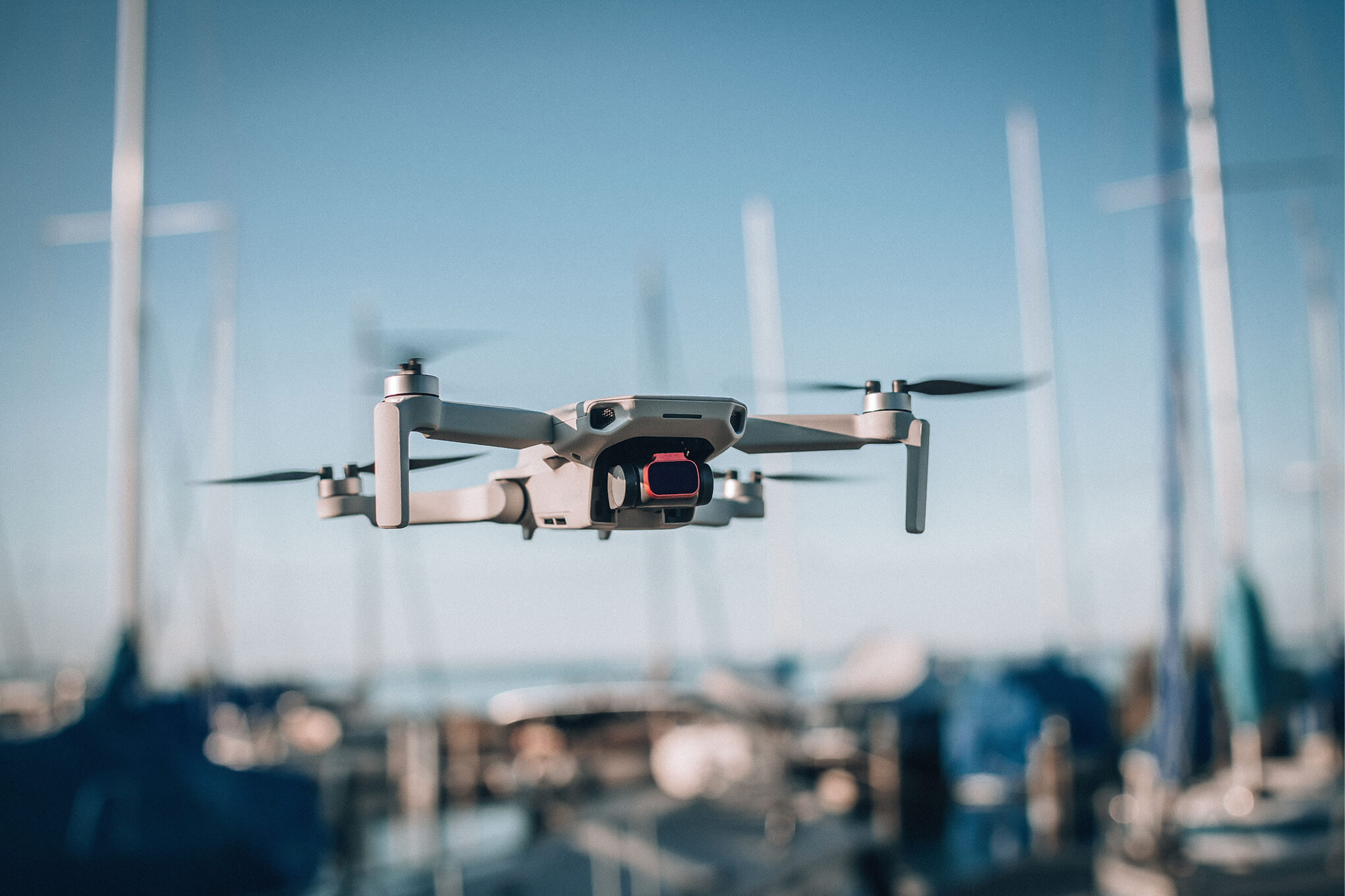 White quadcopter drone flying over a marina with blurred boats and masts in the background.