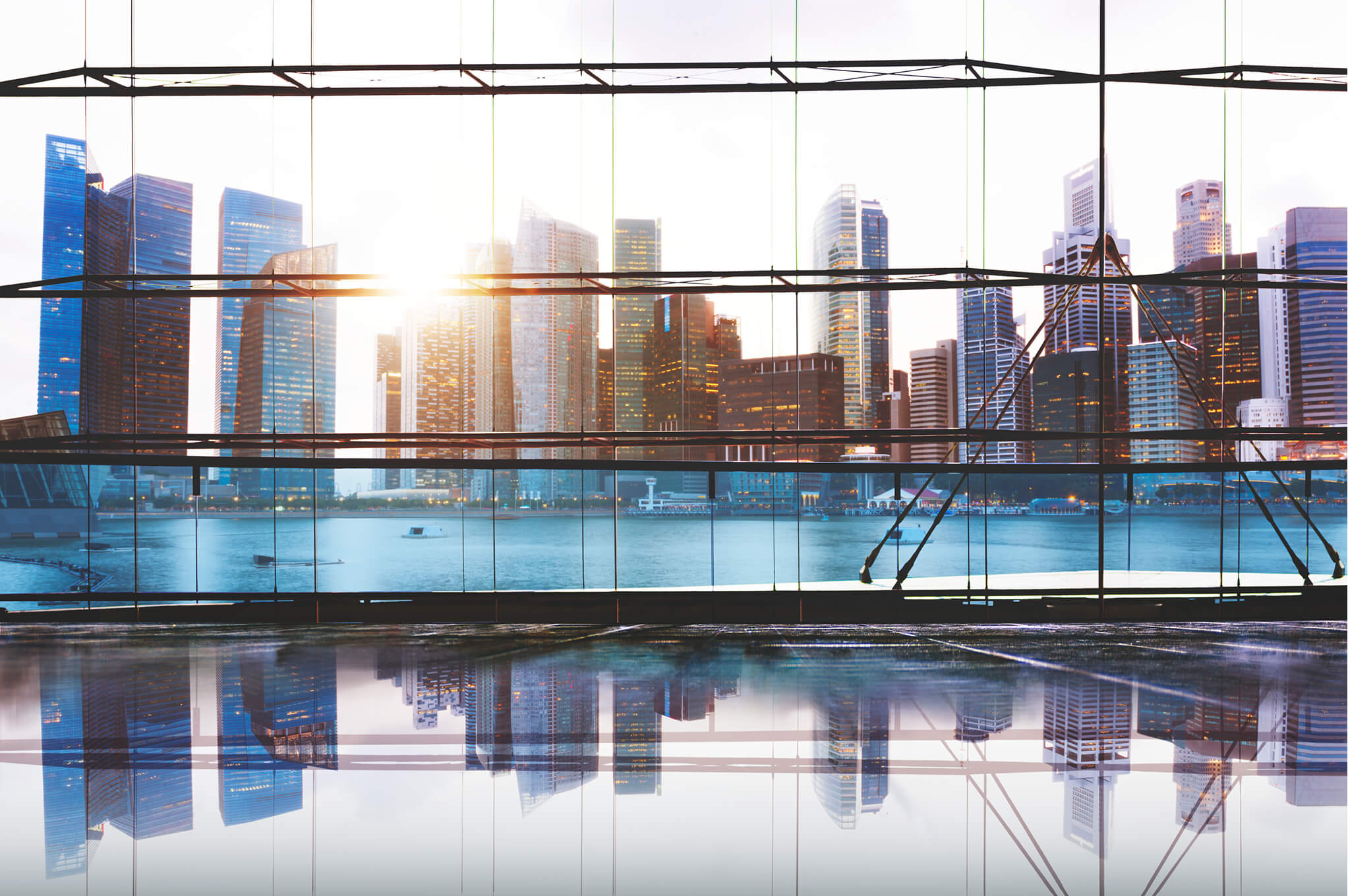 City skyline with modern skyscrapers reflecting on a calm body of water, viewed through large glass windows of a building interior at sunrise or sunset.