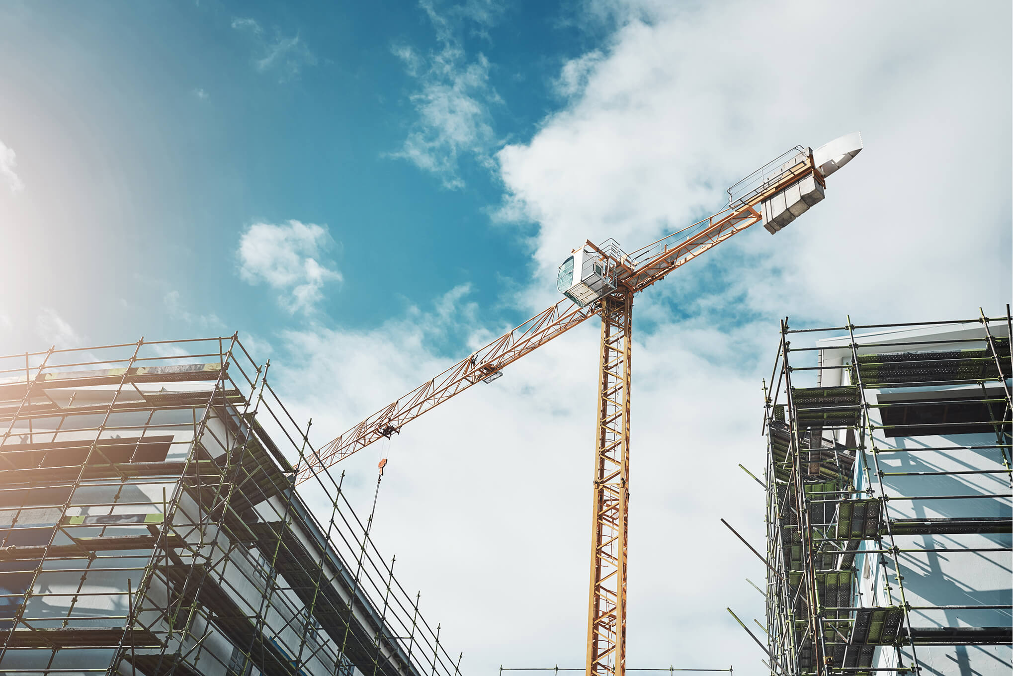 Yellow tower crane between two buildings under construction with scaffolding and a partly cloudy blue sky background.