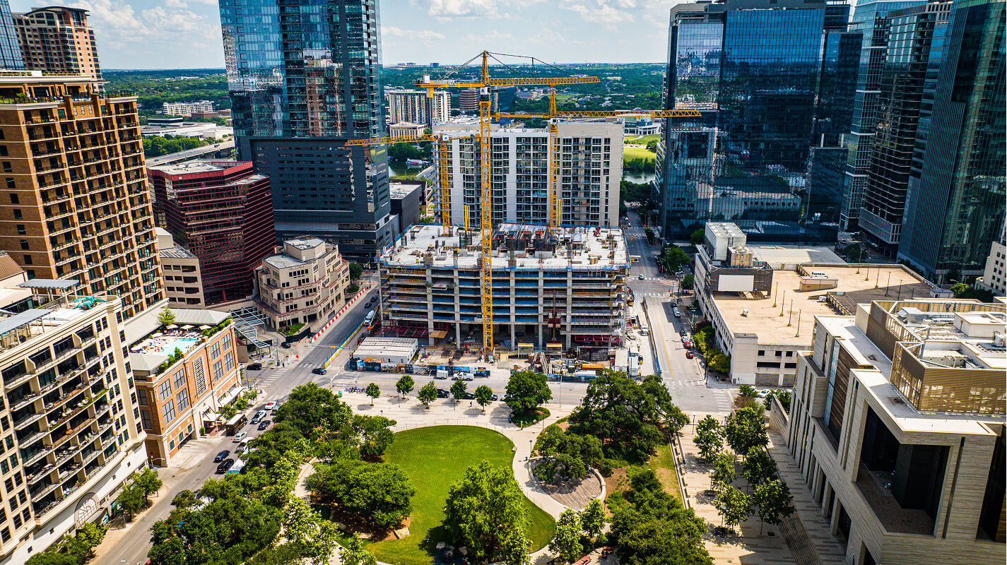 Aerial view of an urban park with green grass and trees in the foreground, surrounded by tall buildings and a construction site with cranes in the background under a partly cloudy sky.