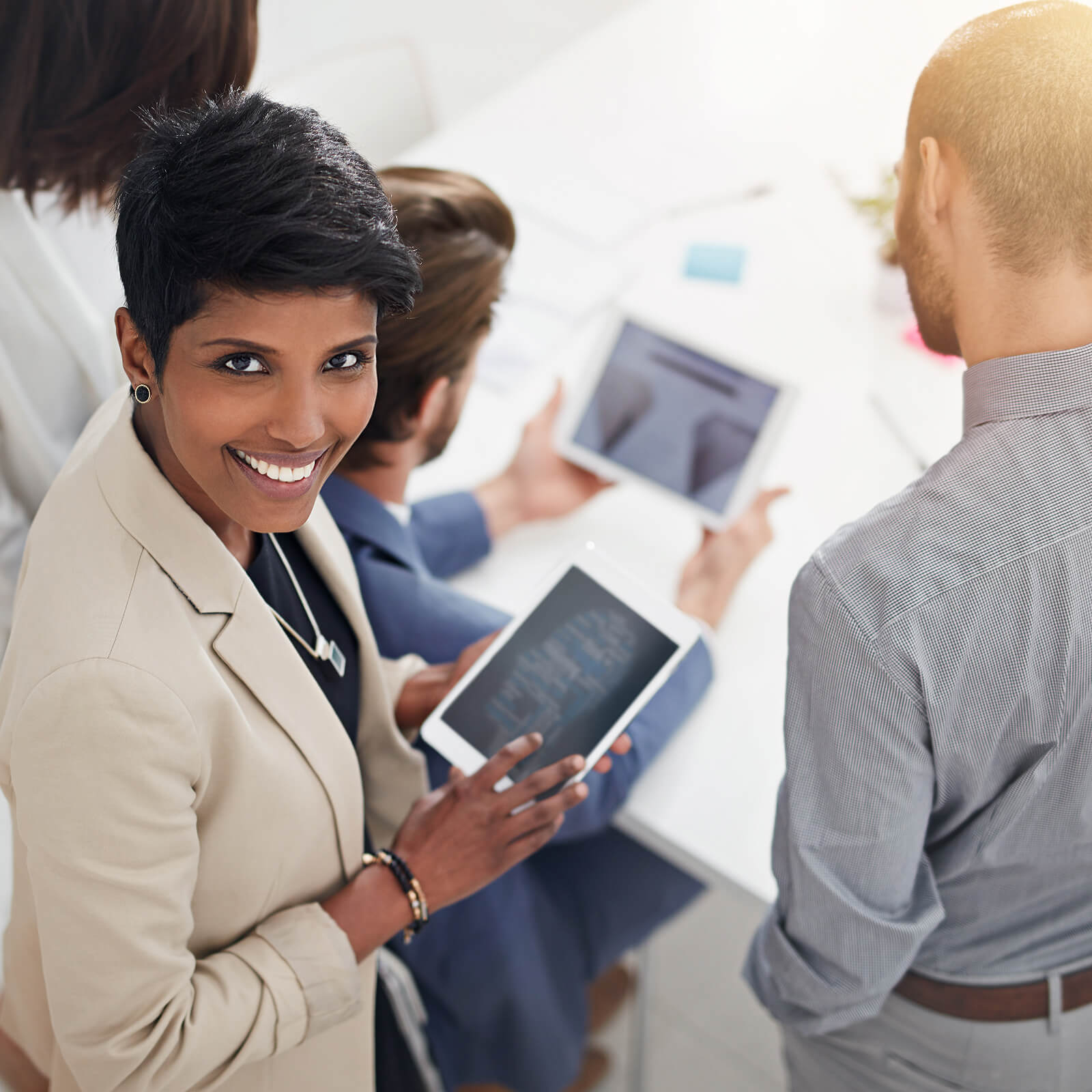 Smiling businesswoman in beige blazer holding a tablet, with three colleagues around a table using tablets in a bright office setting.