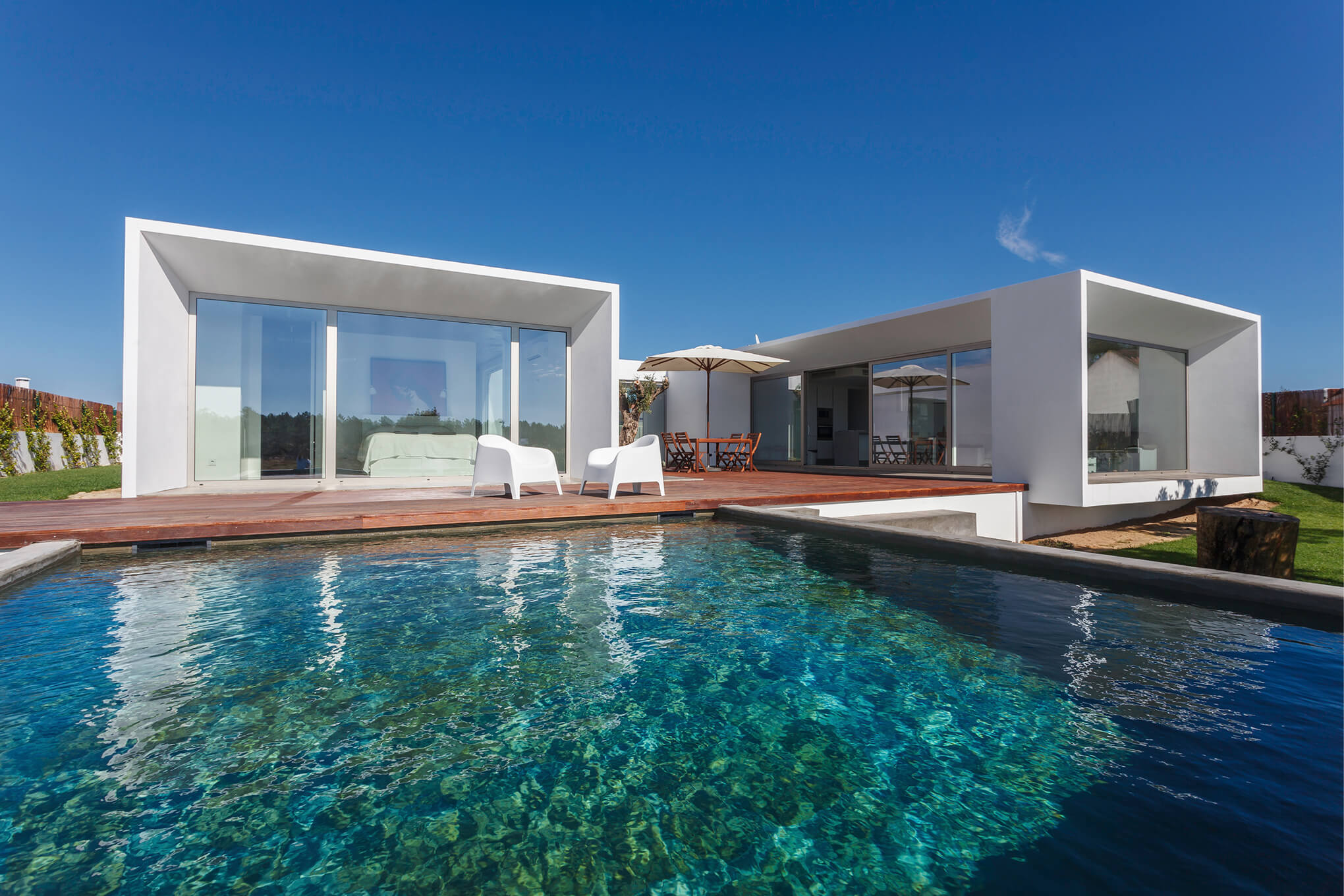 Modern white house with large glass sliding doors, a wooden deck with white chairs and a table under an umbrella, beside a clear blue swimming pool under a bright blue sky.
