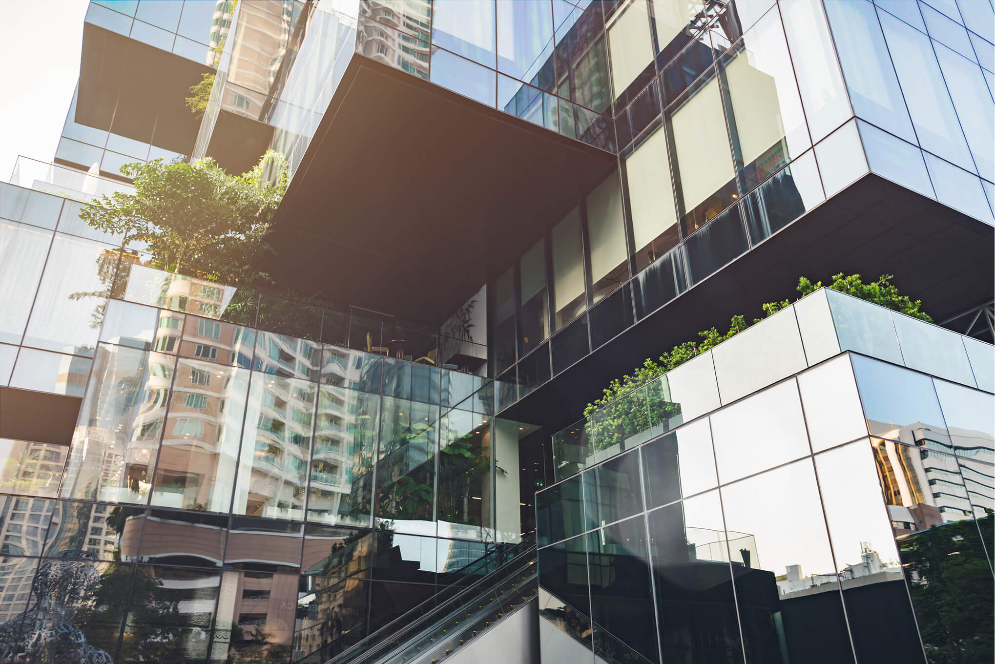 Modern building exterior with large reflective glass panels showing urban buildings reflections, an outdoor staircase, and greenery on balconies.