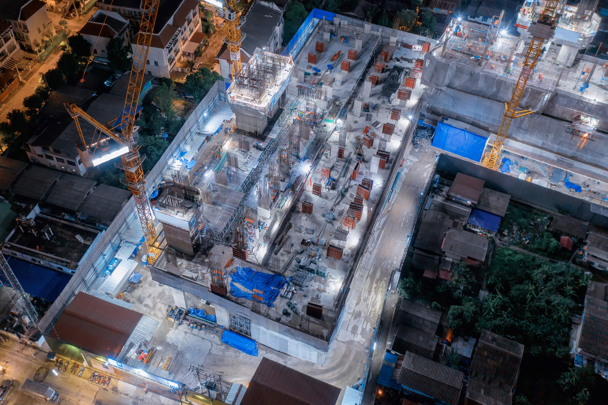 Aerial night view of a large construction site with cranes, concrete structures, scaffolding, and workers illuminated by bright lights, surrounded by residential buildings and roads.