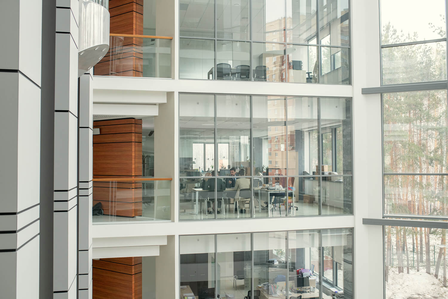 Modern office building interior with large glass windows showing multiple floors and a meeting room where people sit around a table discussing.