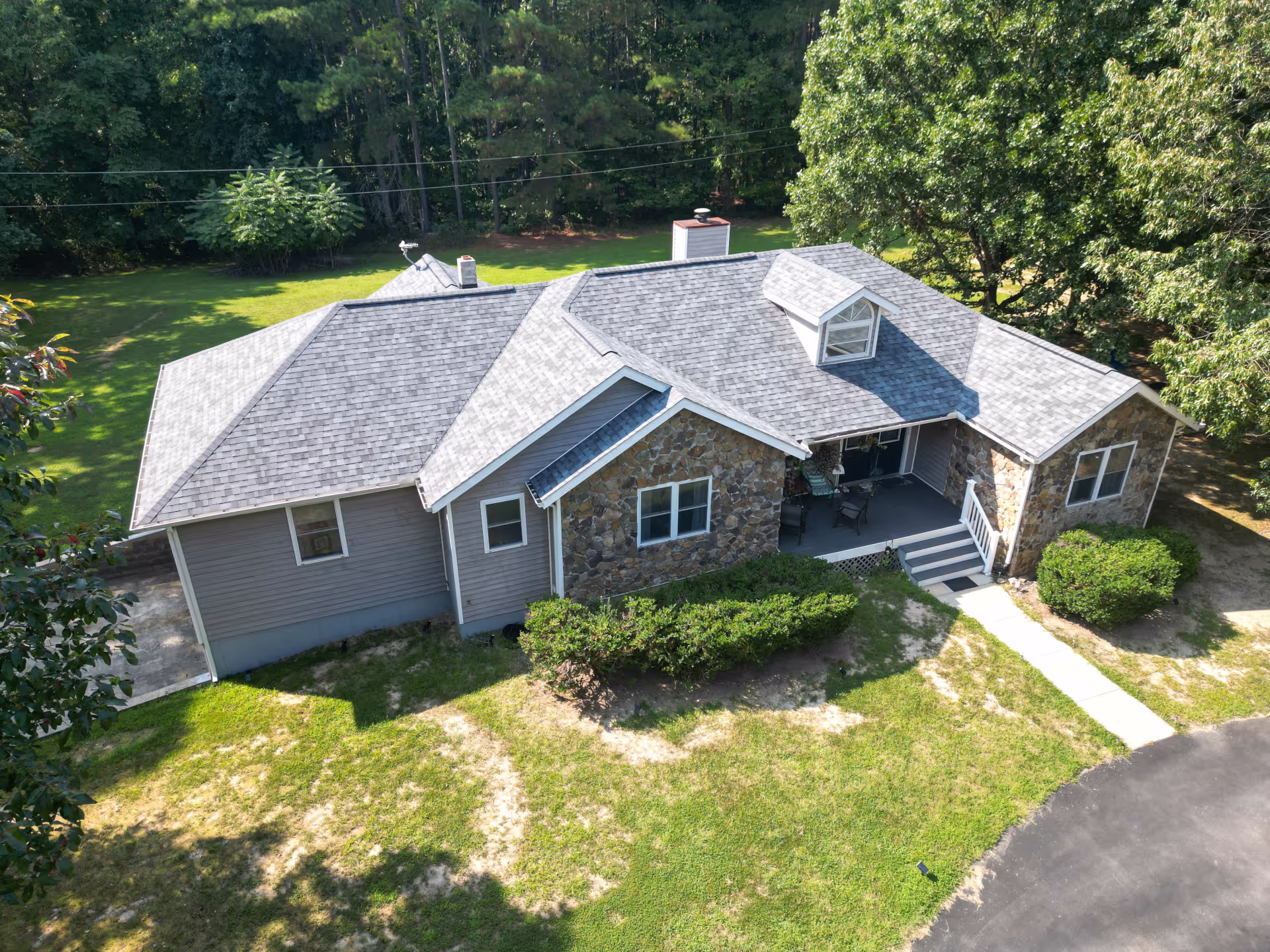 Aerial view of a single-story house with gray roof, stone and gray siding exterior, surrounded by green lawn and trees.