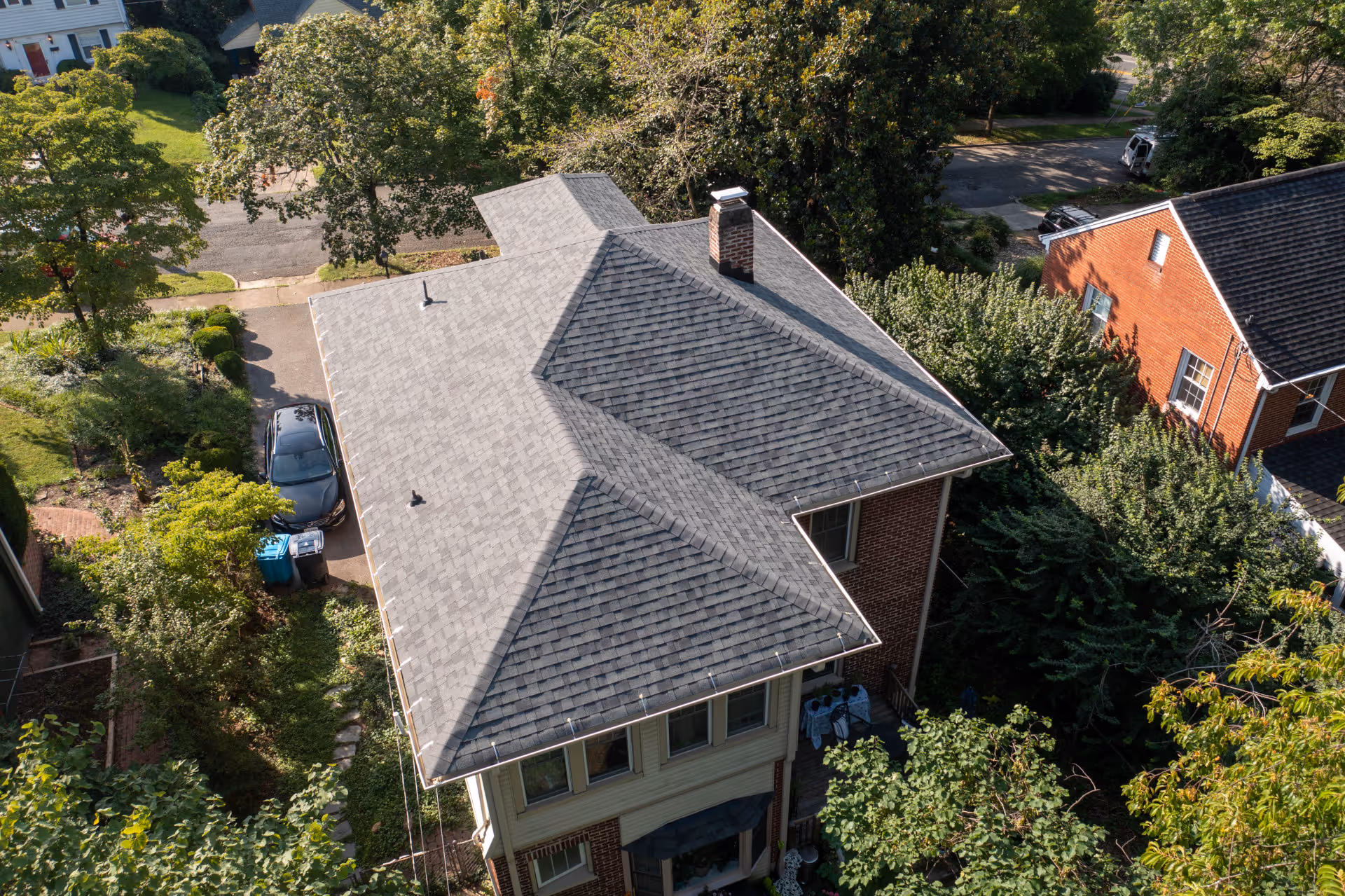 Aerial view of a two-story brick house with a grey shingle roof surrounded by trees and neighboring homes.