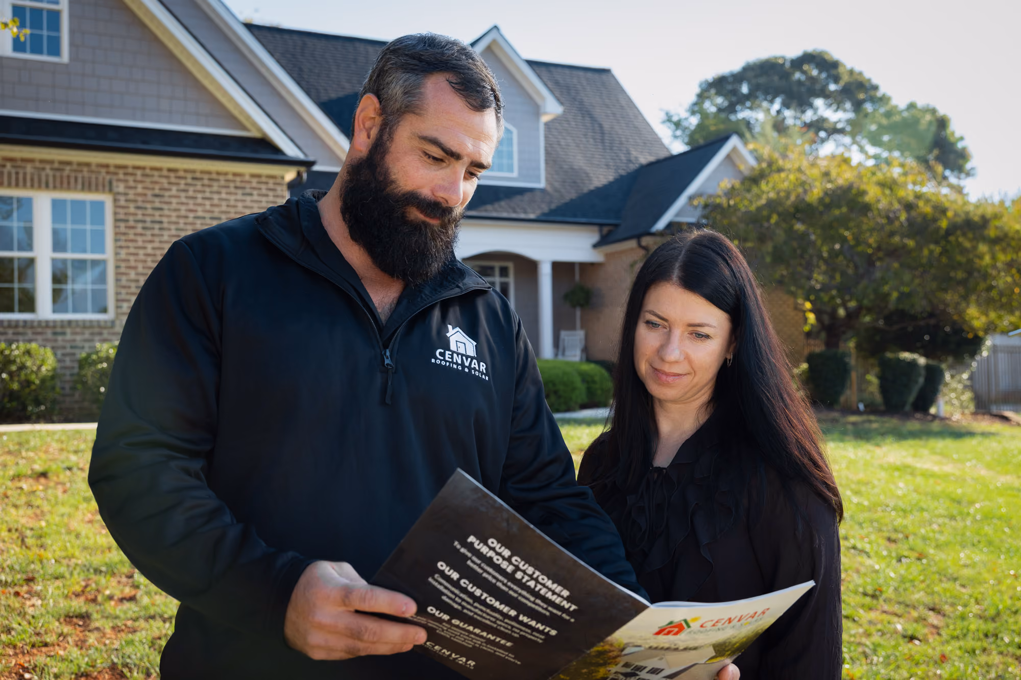  A man and a woman outdoors reviewing a brochure or catalog, with a house and trees in the background.