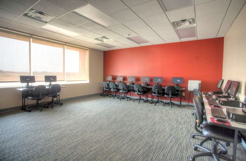 Empty computer lab with multiple workstations lining a bright red accent wall and large windows.