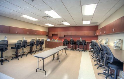An empty classroom with stools along the walls, a long table in the center, and microscopes on a counter.