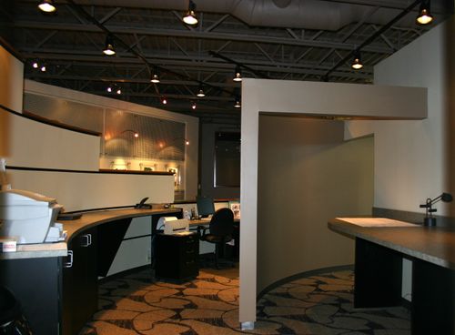 A room featuring a large white wall and a black and white counter, accompanied by various office furniture including chairs and a desk.