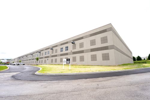 A building is situated alongside a curved road, surrounded by grass and trees, with a clear sky above.
