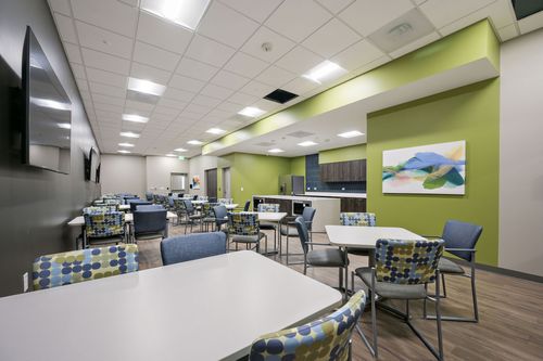 A room featuring multiple tables and chairs, with a wall-mounted whiteboard, ceiling lights, and a clean floor.