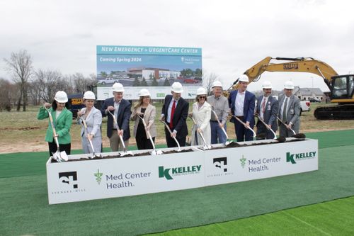 A group of people in hard hats holding shovels stands outdoors on grass, with a sign reading "NEW EMERGENCY + URGENT CARE CENTER Coming Spring 2024" visible.