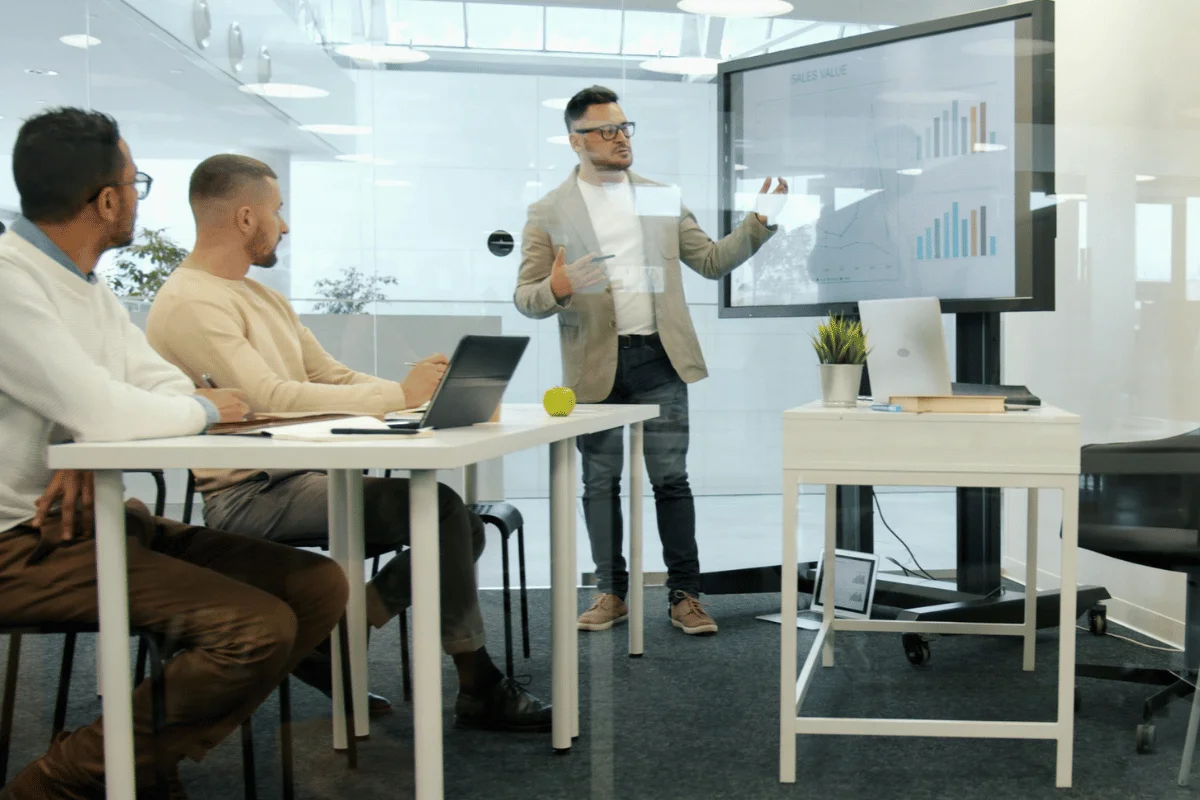A man presents sales graphs on a screen to two seated colleagues in a modern, glass-walled office. The setting is professional and focused.
