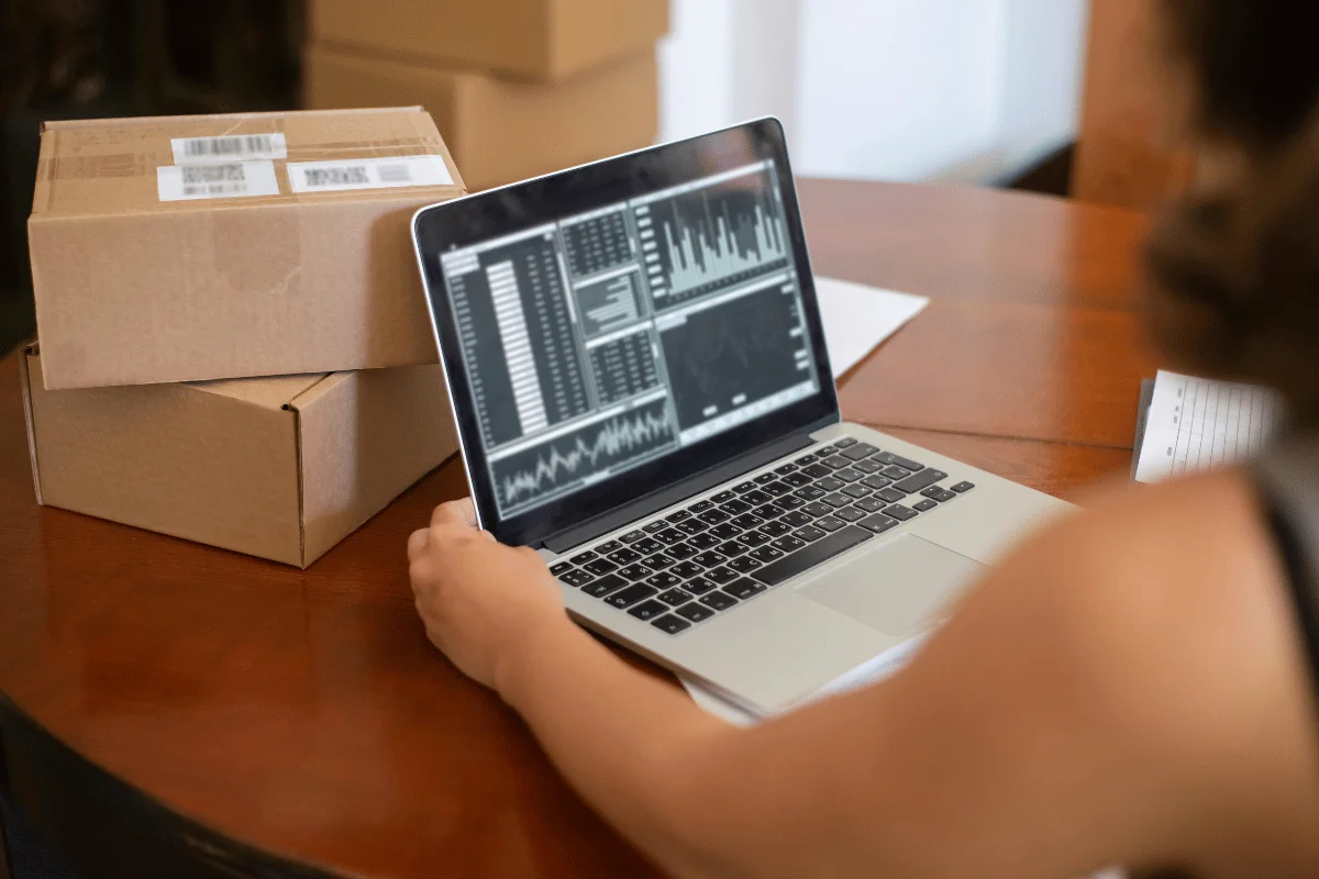 A person works at a wooden table on a laptop displaying financial graphs and data. Nearby, stacked cardboard boxes suggest business or shipping tasks.
