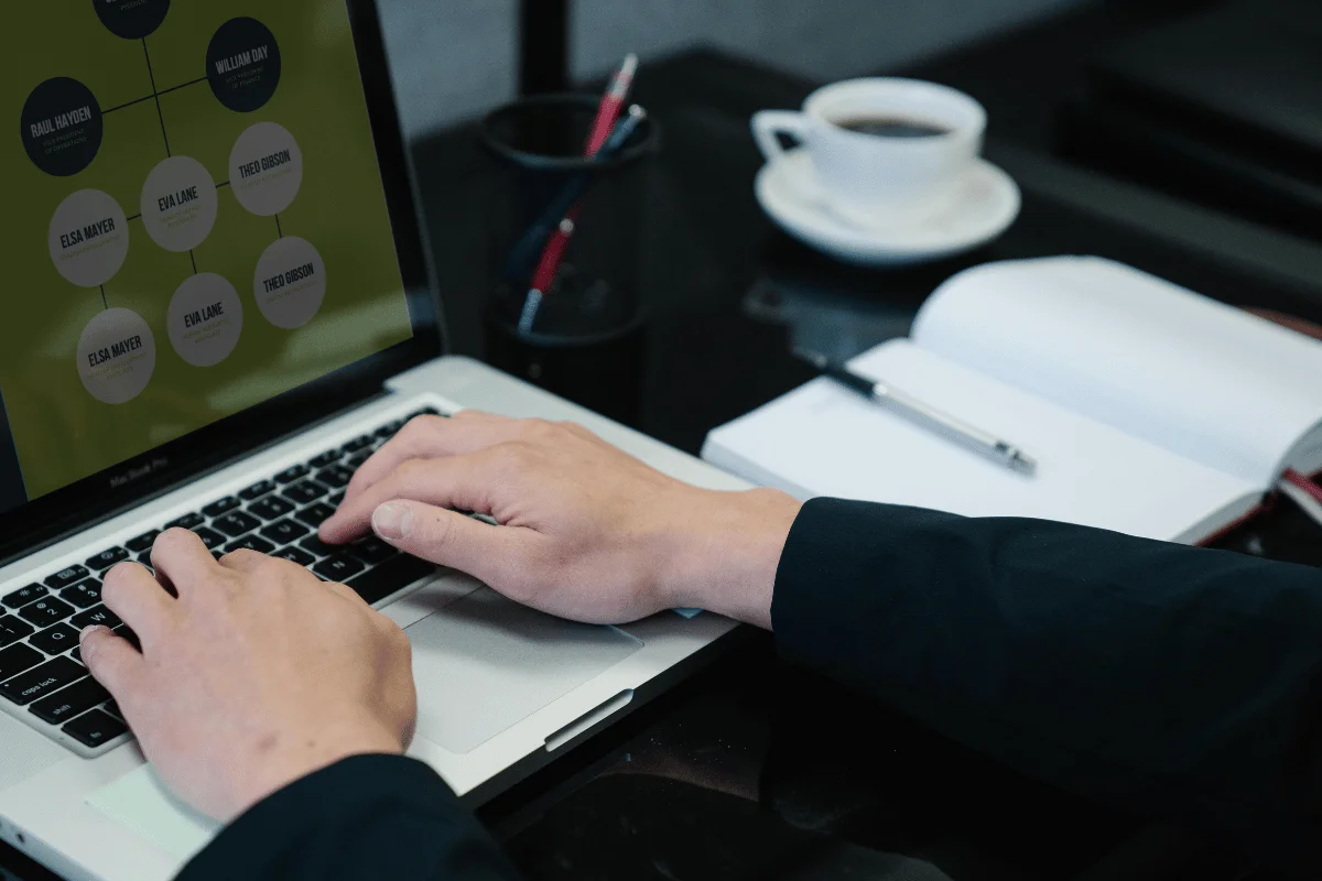 Hands on a laptop keyboard displaying a flowchart. Nearby, an open notebook with a pen and a cup of coffee. The scene conveys focus and productivity.