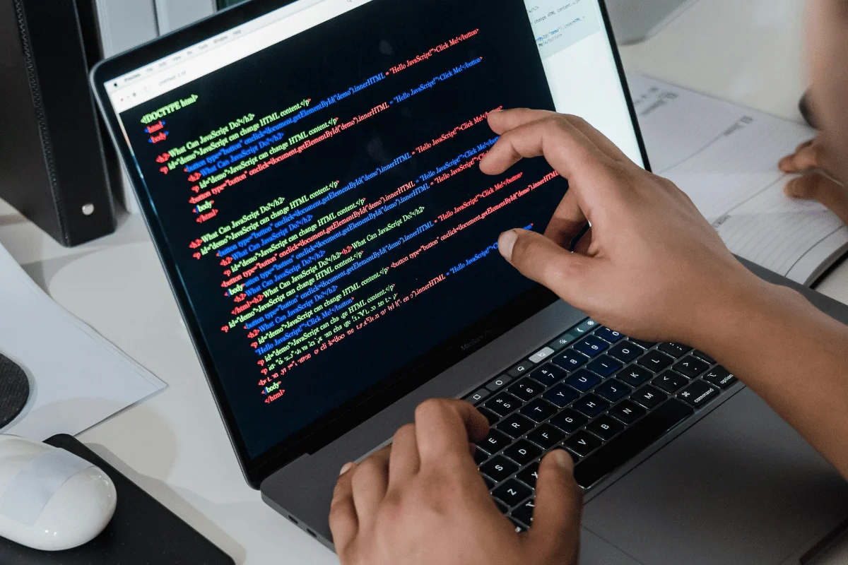 Hands typing code on a laptop displaying colorful HTML text, indicating programming activity. The setting suggests a focused, productive workspace.