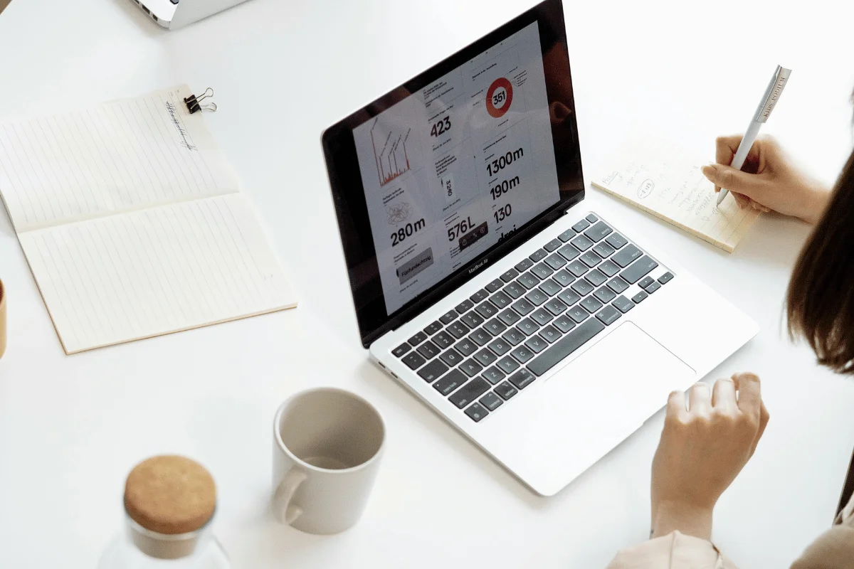 A laptop displaying charts and graphs is on a white desk. A person writes in a notebook beside it. A coffee cup and notepad are nearby, suggesting a workspace setting.