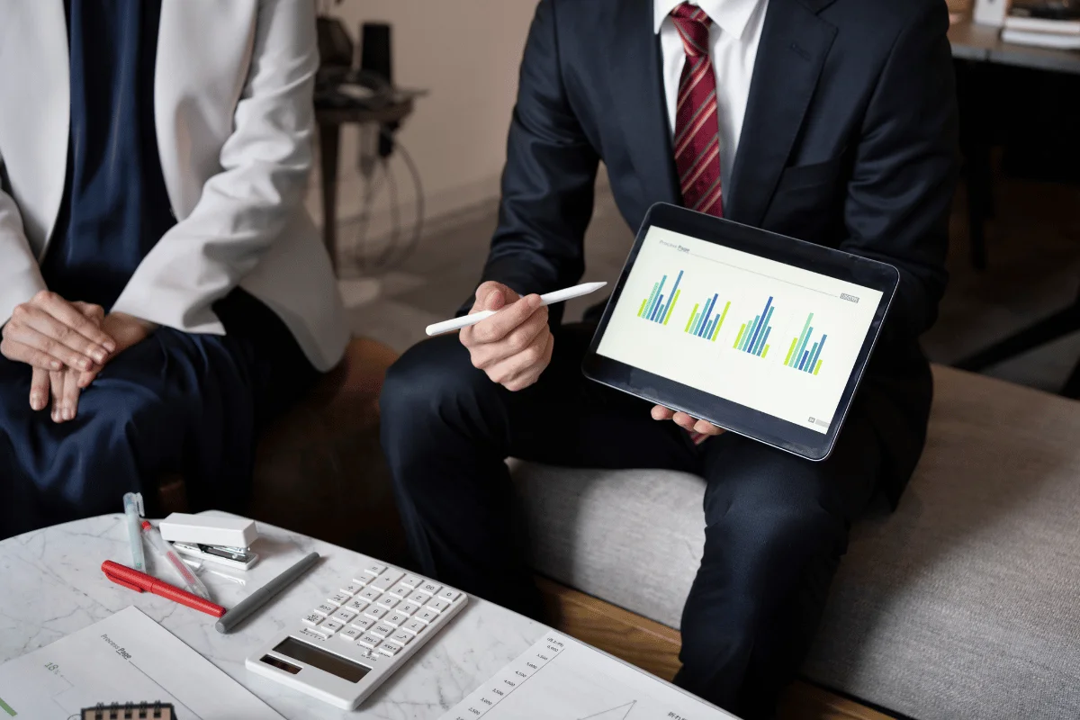 A business professional in a suit presents bar charts on a tablet during a meeting. A calculator and documents are on the table, indicating financial discussion.
