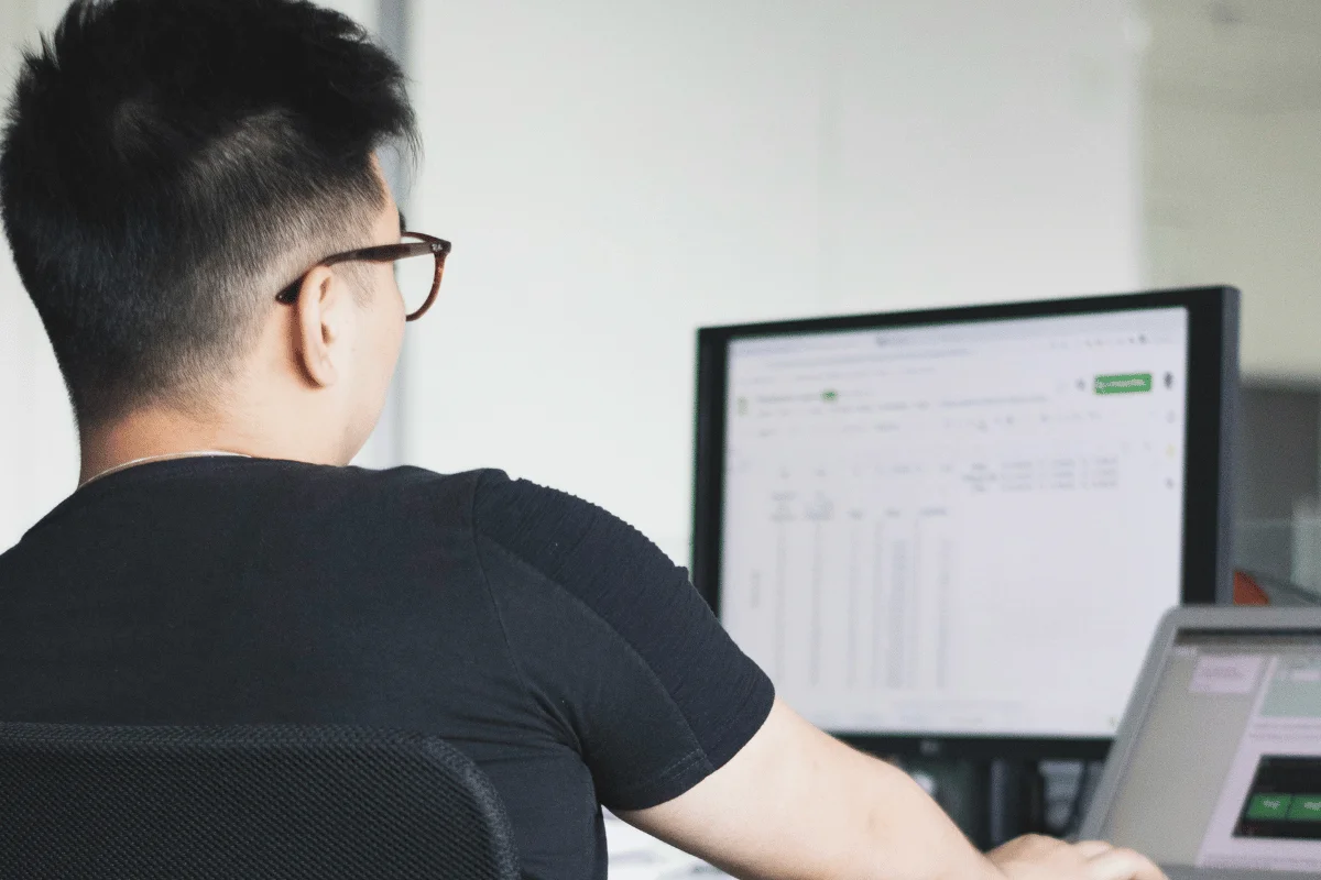 A person in glasses, wearing a black shirt, works at a desk with computers displaying spreadsheets. The setting suggests focus and productivity.