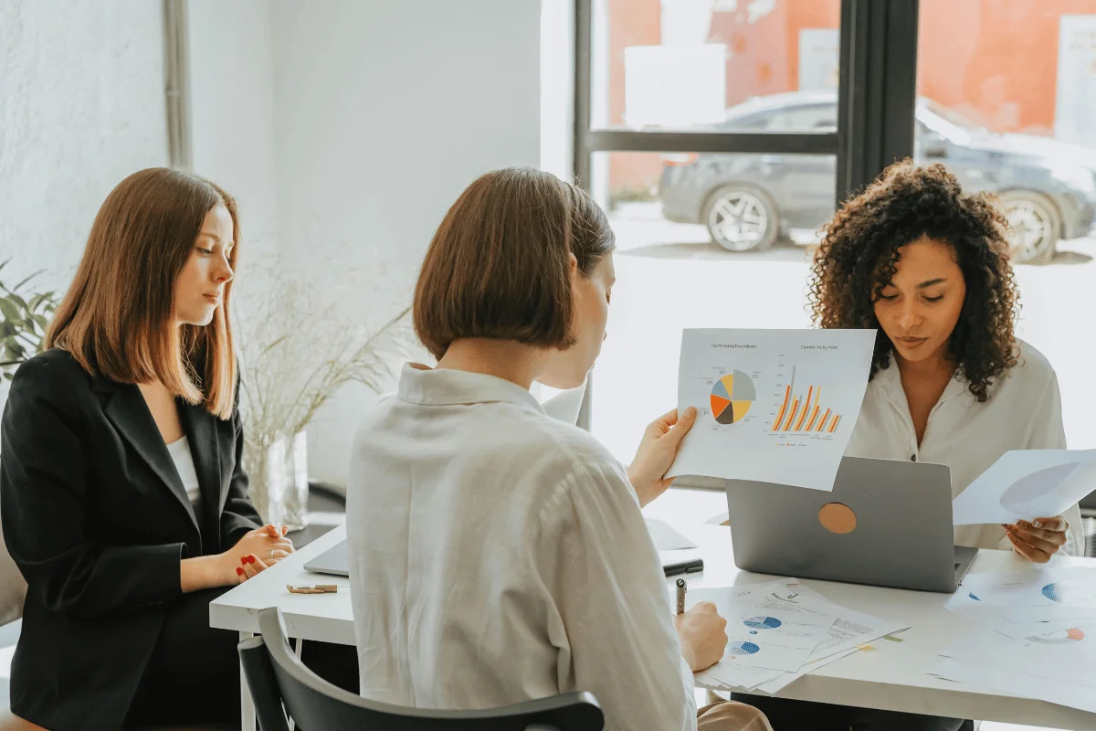 Three women sit at a table in a bright office, reviewing documents with charts and graphs. One uses a laptop, creating a focused, collaborative atmosphere.