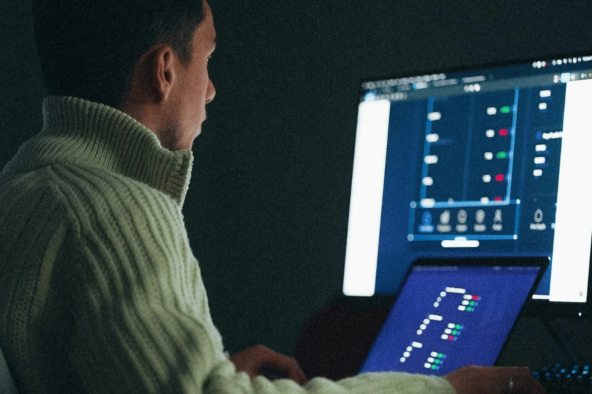 A person in a white sweater focuses on a computer screen displaying complex charts and data in a dimly lit room, conveying concentration and analysis.