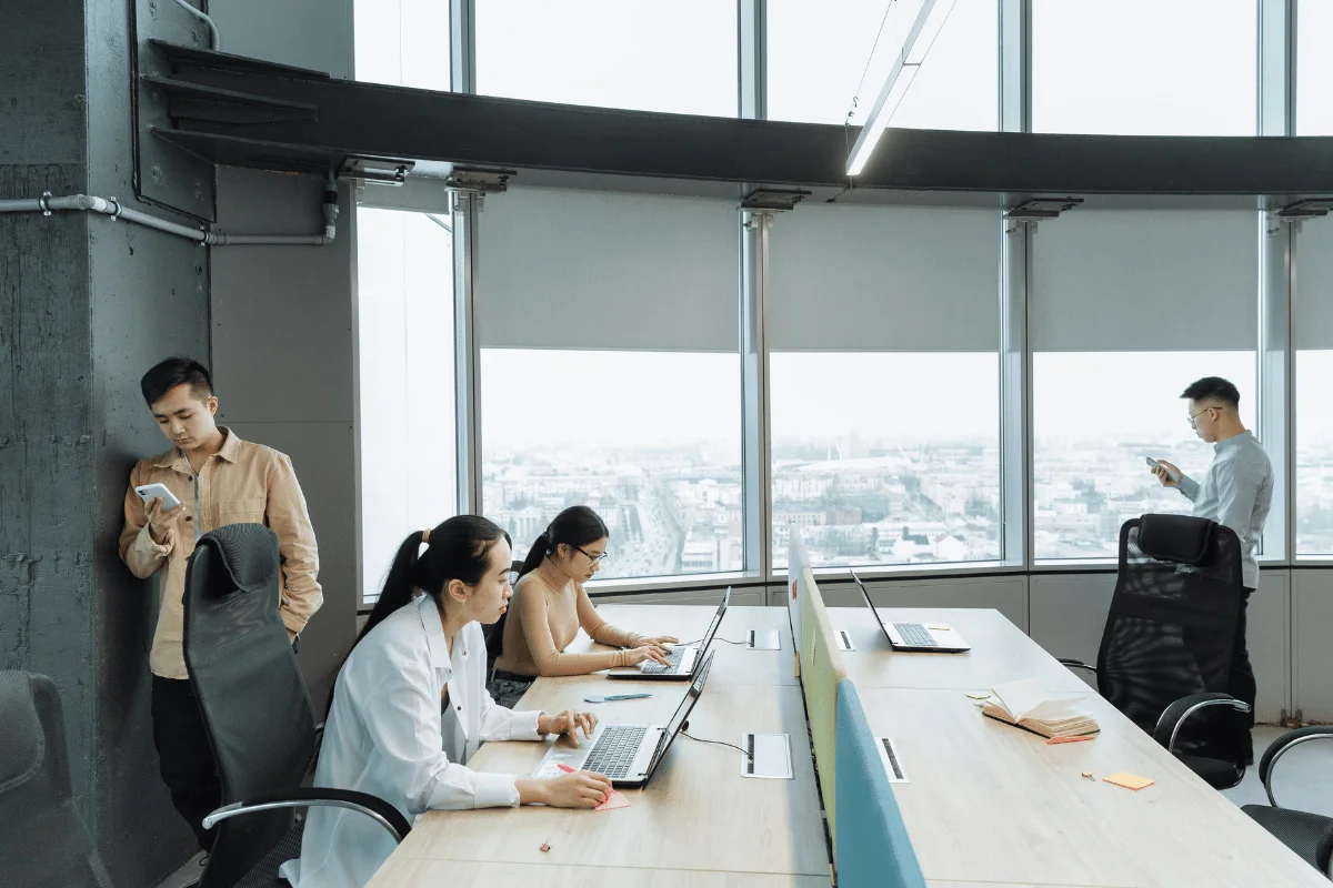 A modern office with large windows showing a cityscape. Two people work on laptops at a long desk, while two others stand by the windows using phones.