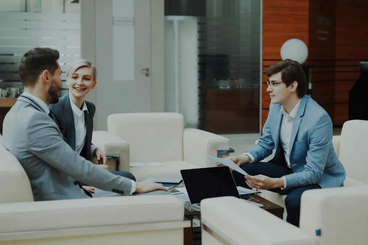 Three people in a modern office lounge, sitting on cream-colored chairs. They are discussing with open laptops and papers, appearing engaged and cheerful.