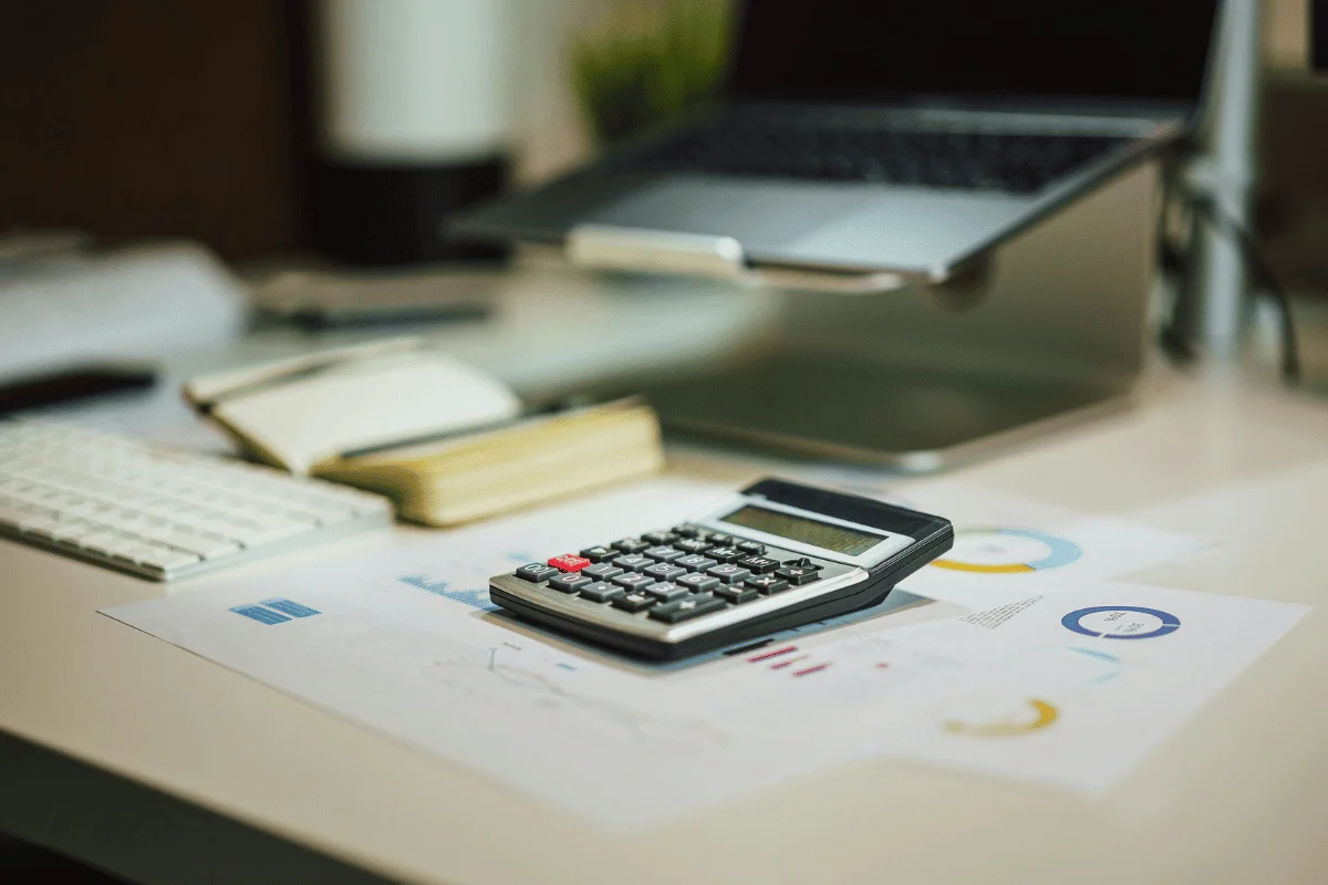 A calculator rests on printed charts and graphs, with a laptop and notebook visible in a tidy workspace.