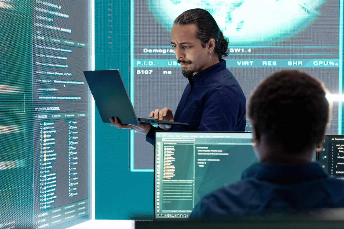 A man in a blue shirt works on a laptop in a high-tech room with glowing digital screens displaying complex data and code, creating an intense, focused atmosphere.