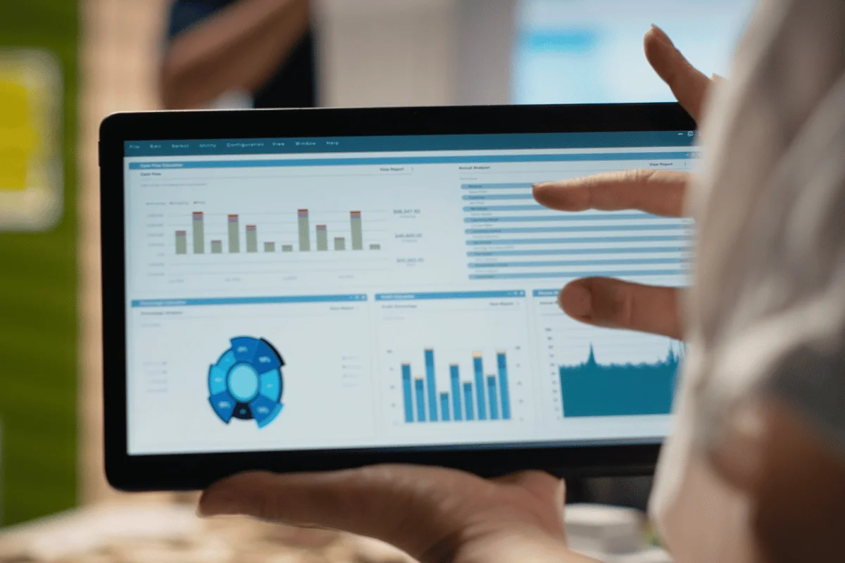 Close-up of a person using a tablet displaying data charts and graphs. A hand points at the screen, suggesting analysis in a work or tech setting.
