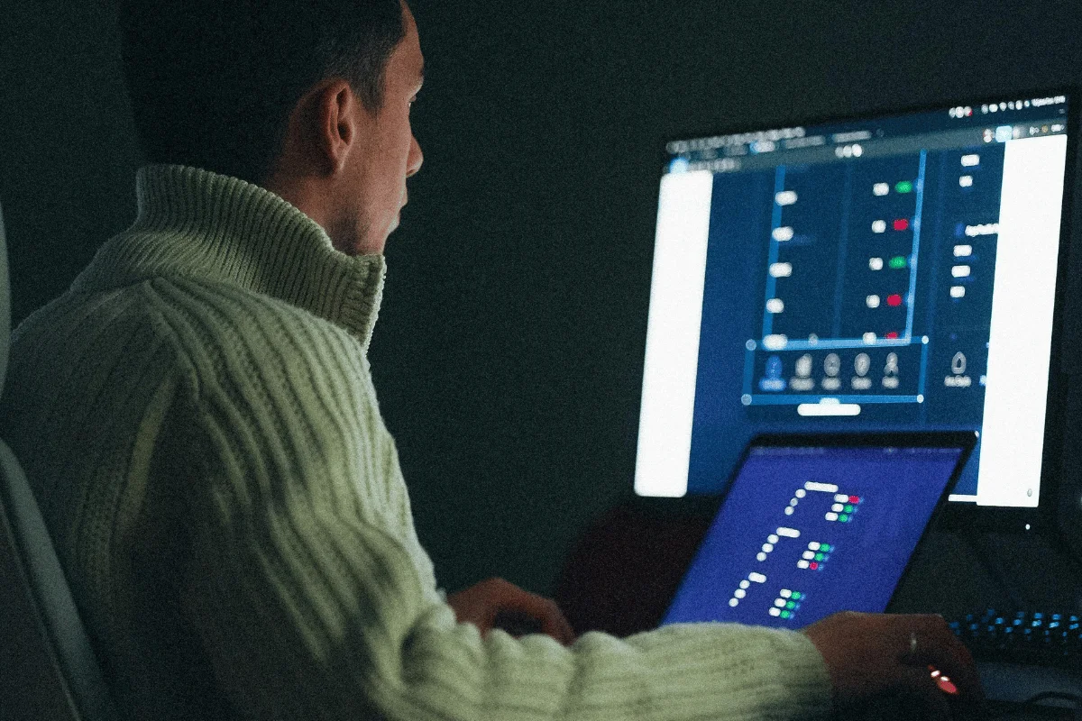 A person in a cozy sweater is seated at a desk, working on a laptop with a second monitor displaying data analytics.