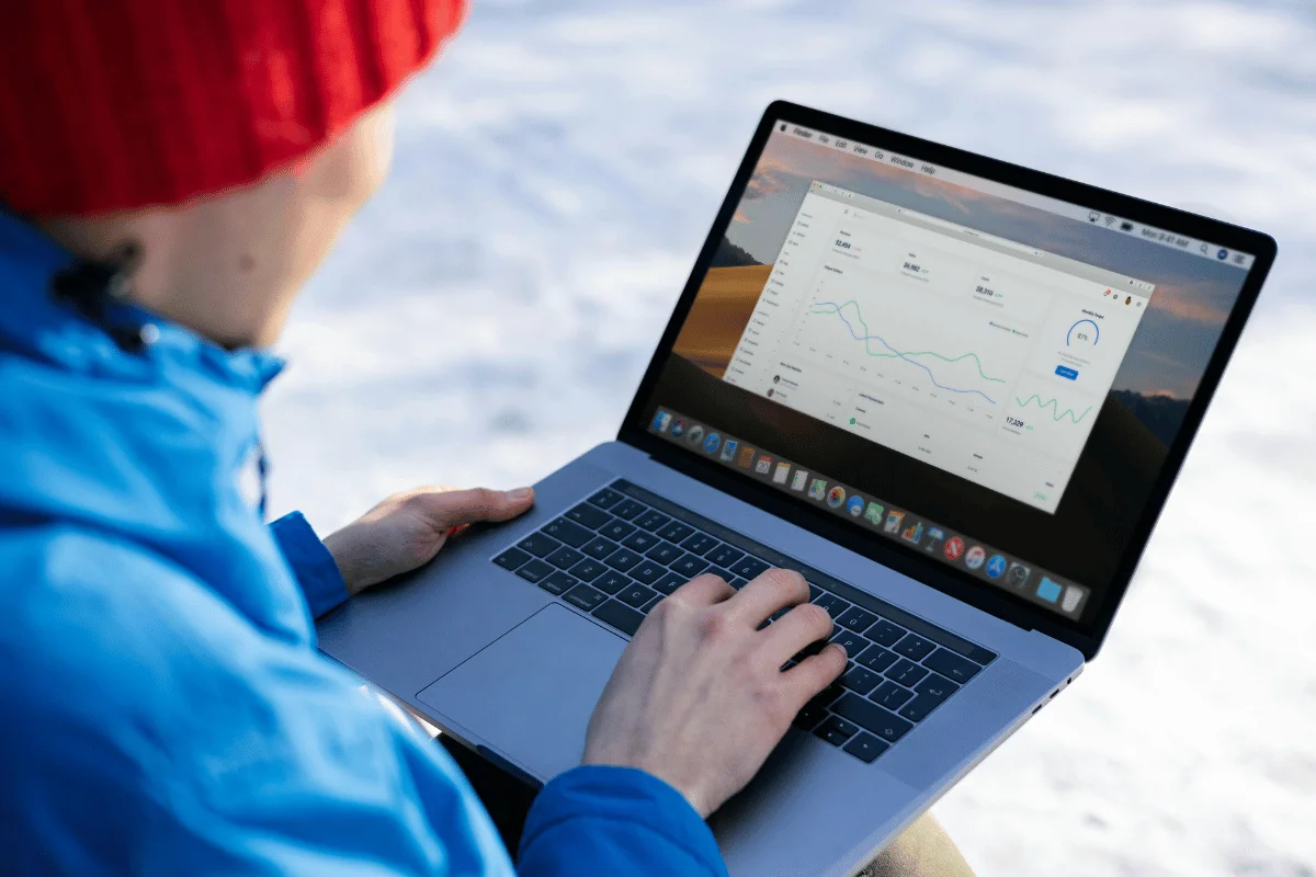 Person in a red hat and blue jacket uses a laptop outdoors on snow. The screen shows a graph and data, conveying focus and analysis in a cold setting.