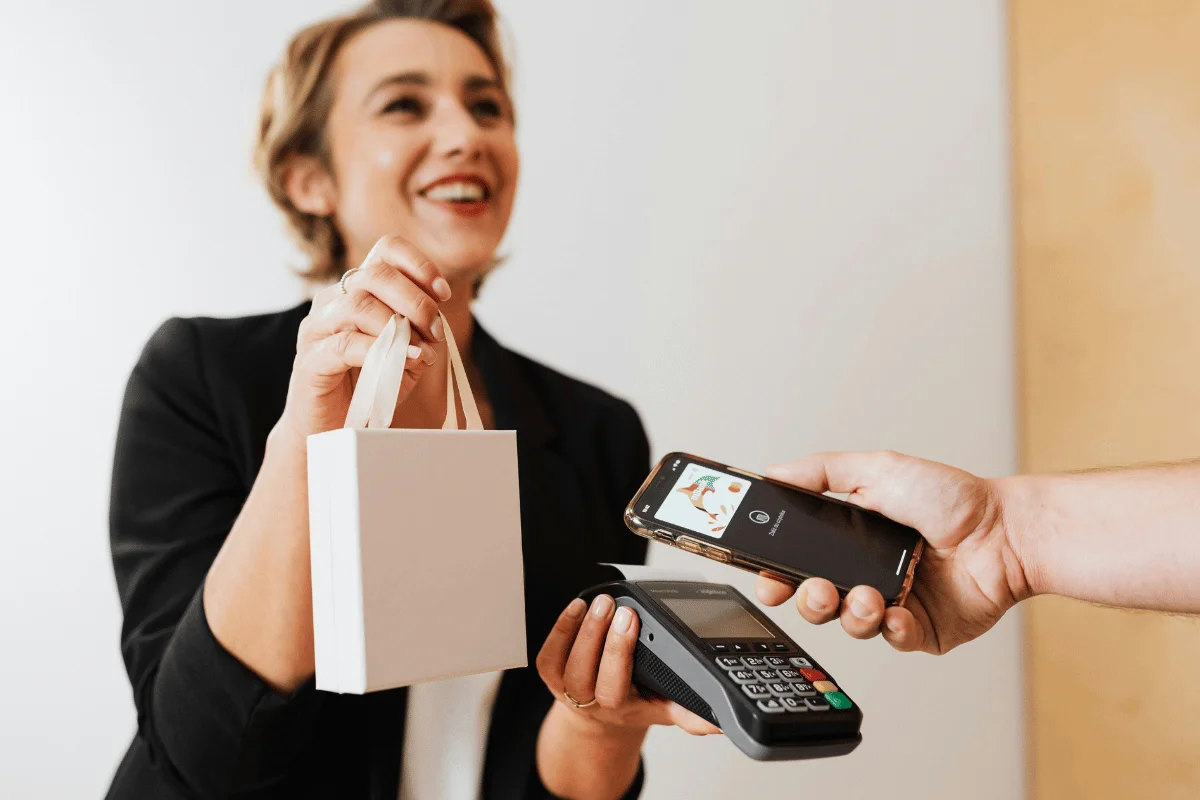 A person holds a small white bag while using a smartphone to make a contactless payment on a terminal.