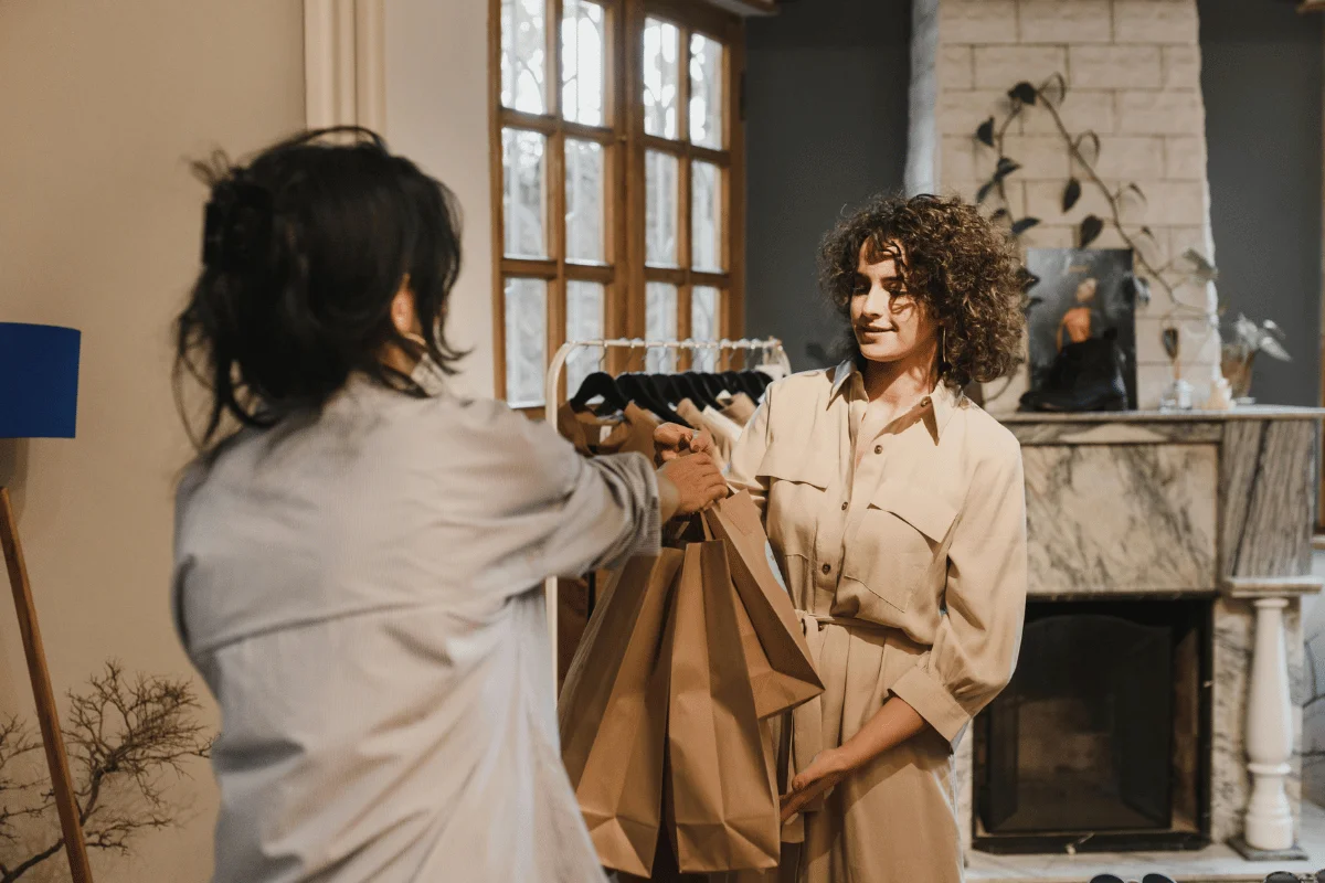 Two women exchanging paper shopping bags in a cozy living room with a stone fireplace and large window.