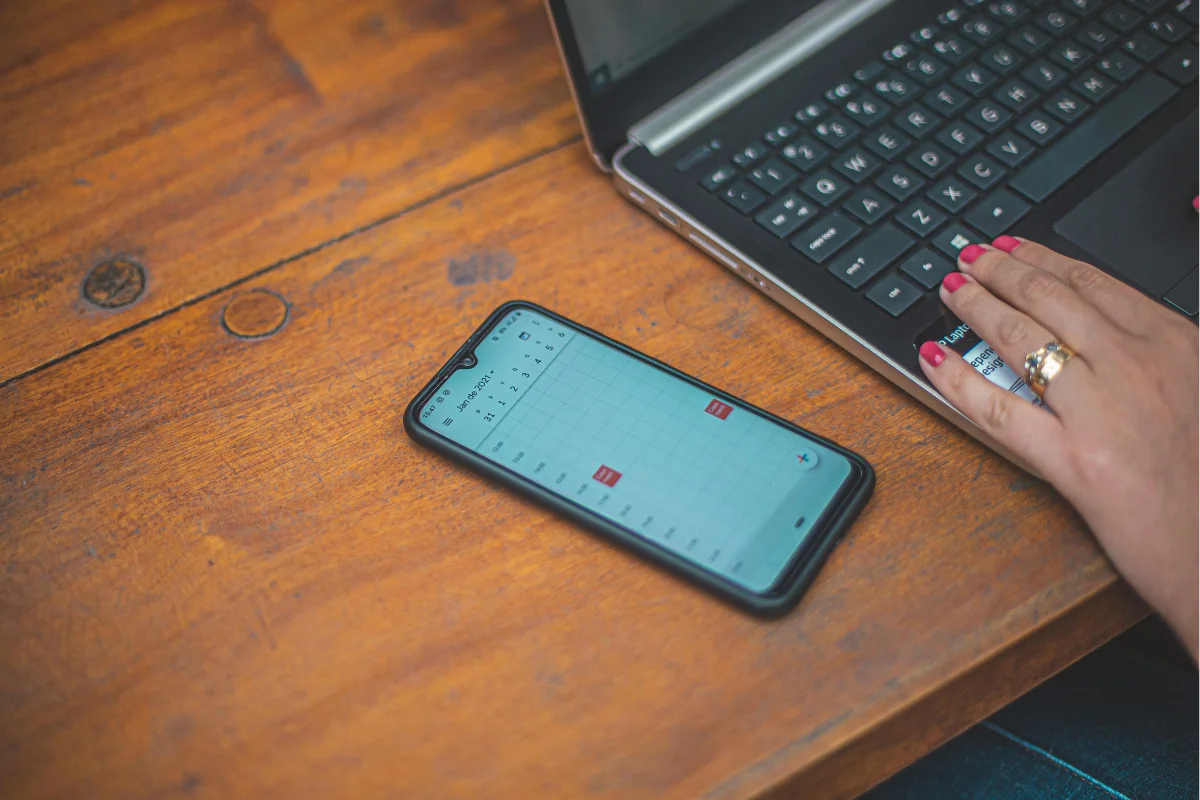 A smartphone displaying a calendar app sits on a wooden table next to a laptop keyboard, with a hand resting on the laptop.