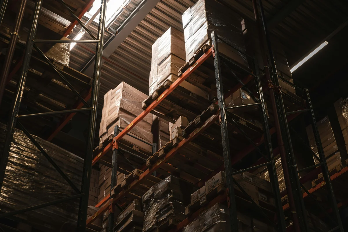 A high-angle view of a warehouse with tall shelving filled with neatly stacked boxes and pallets, illuminated by overhead light.