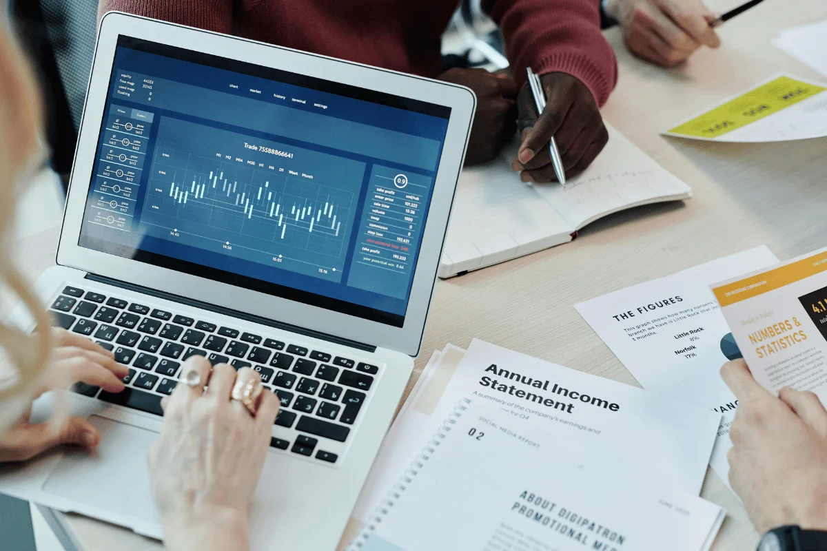 A laptop displaying a financial chart with various data points, surrounded by papers and hands engaged in discussion.