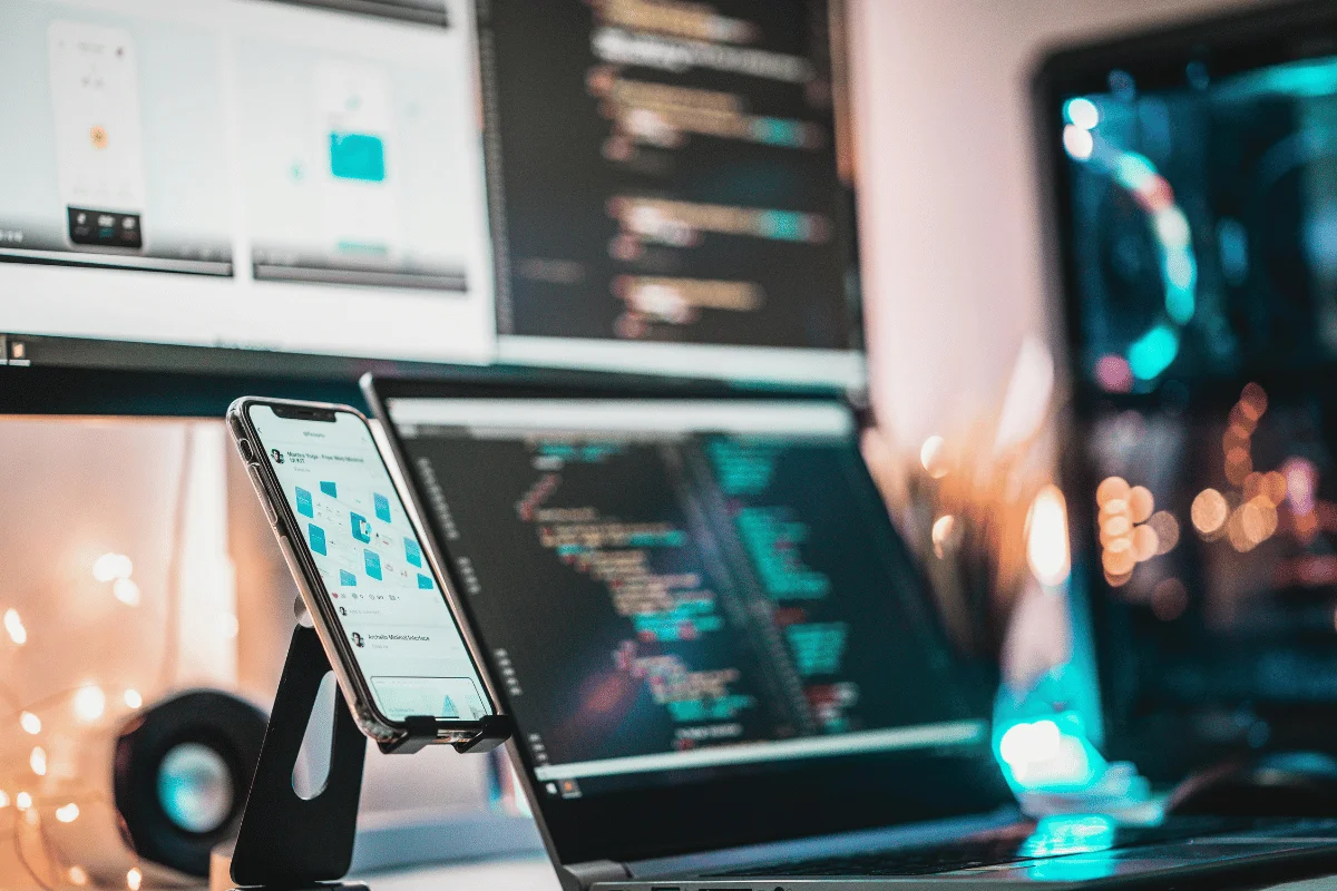 Workspace with a laptop displaying code, a smartphone on a stand showing app design, and blurred monitors with code and graphics in the background.