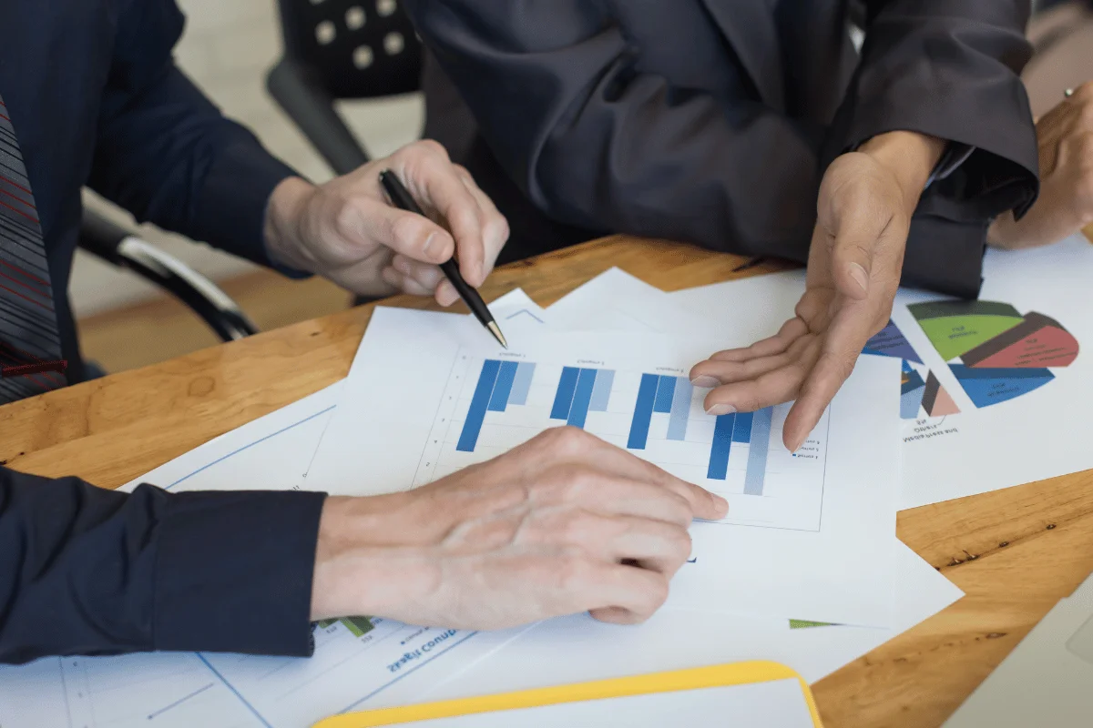 Two people in business attire analyzing printed charts and graphs on a wooden table during a meeting.