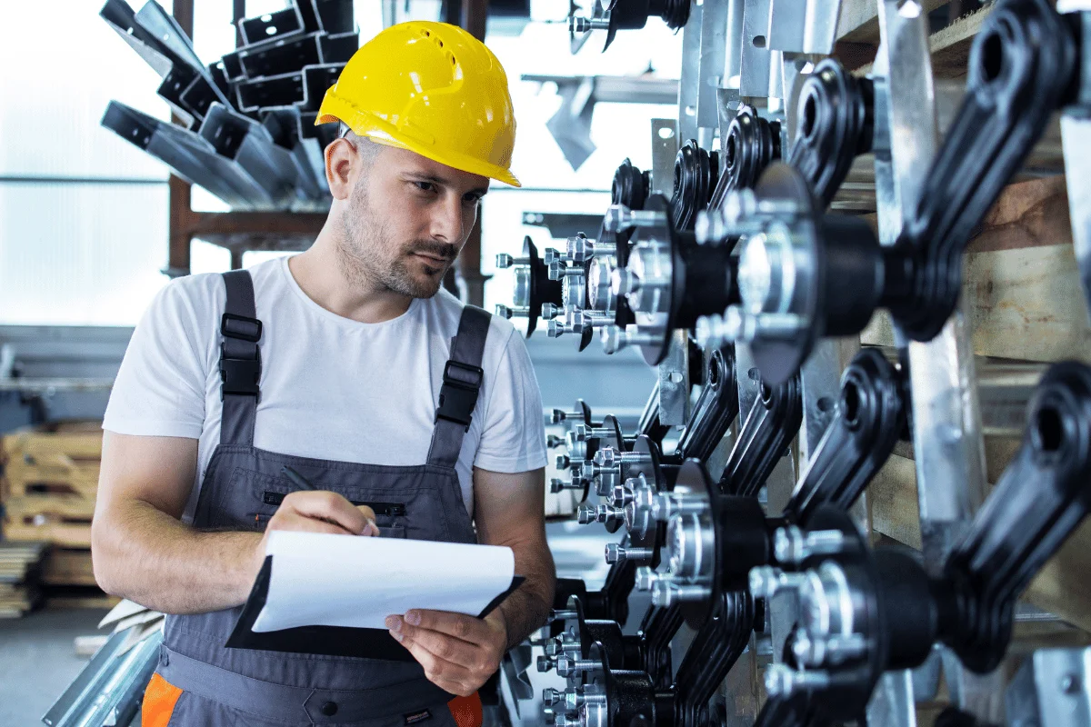 Worker in a yellow helmet and gray overalls inspects and takes notes on mechanical parts in an industrial warehouse.