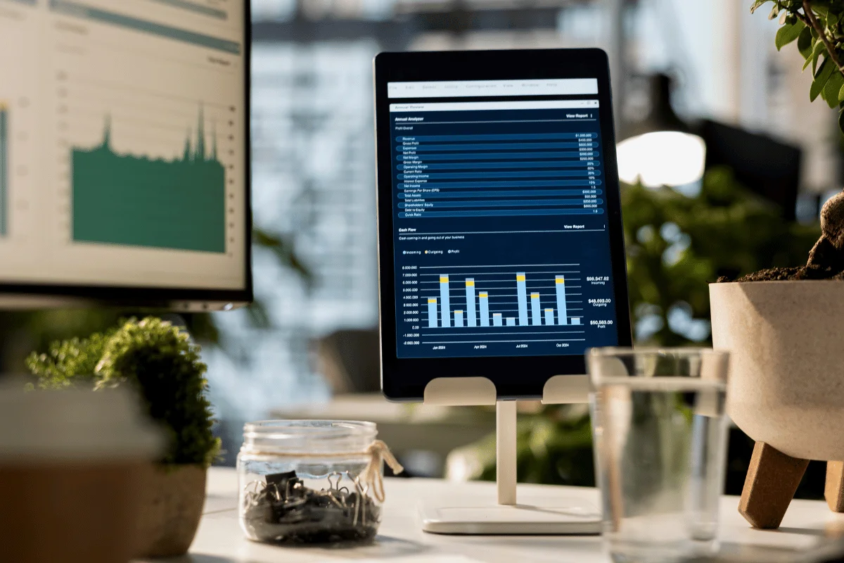 Tablet on a desk displaying bar charts and data; a glass of water, jar, and potted plant nearby.