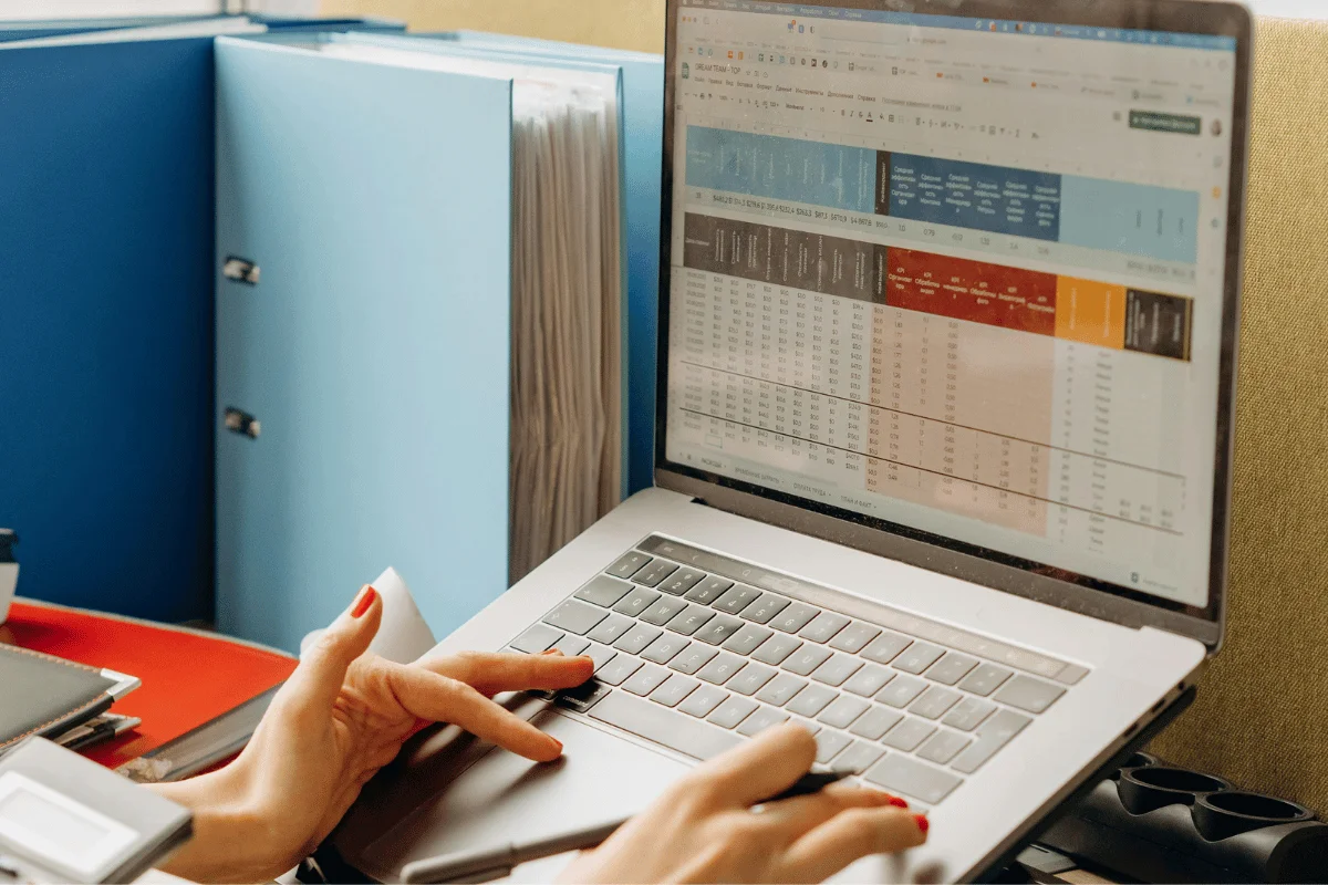Hands typing on laptop while reviewing financial spreadsheet and documents at office desk.