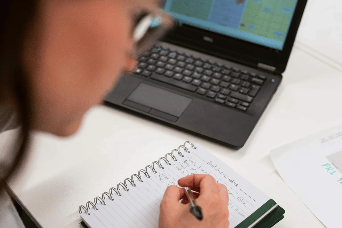 Professional writing notes in a spiral notebook beside a laptop showing a calendar or scheduling dashboard.