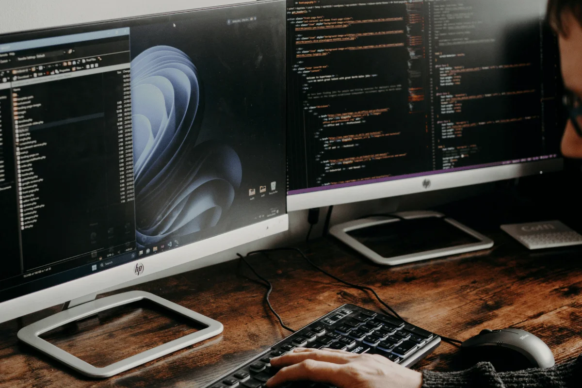 Programmer working at dual monitors displaying code and software development tools on a desk.