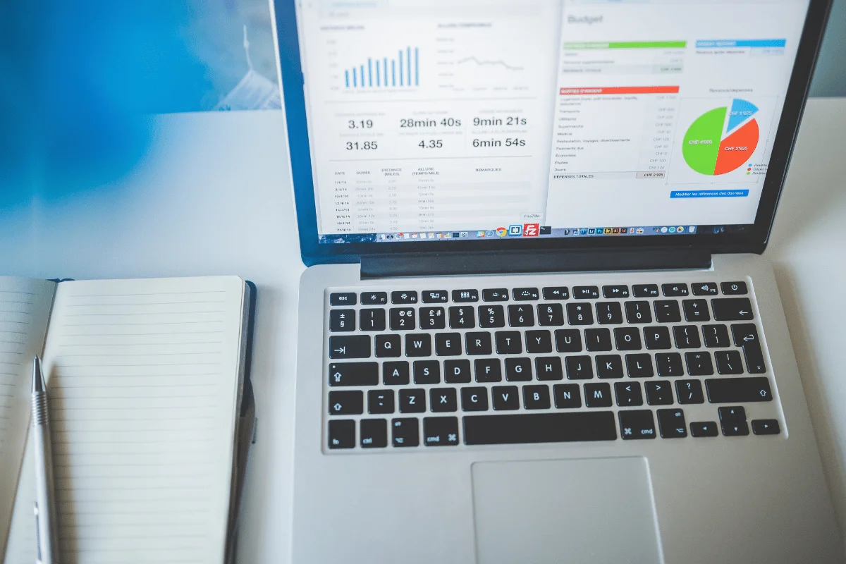 Laptop displaying business dashboards and budget charts beside a notebook and pen on a desk.
