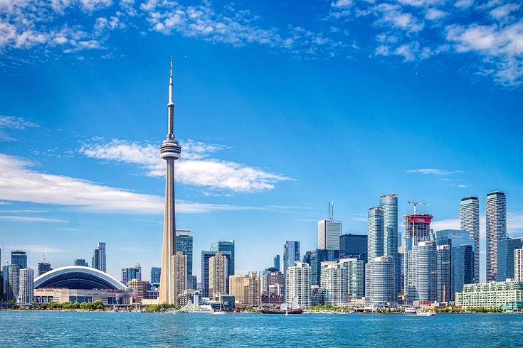 the iconic skyline of Toronto, Ontario, viewed from across Lake Ontario. Dominating the center is the CN Tower, one of Canada’s most recognizable landmarks. Surrounding it are modern high-rise buildings, including office towers and residential condos, reflecting the city’s urban density and architectural growth. To the left of the CN Tower is the Rogers Centre, a large stadium with a retractable roof. The foreground features calm water that mirrors the skyline, while the sky above is mostly clear with a few scattered clouds, adding to the scenic and vibrant feel of the image.
